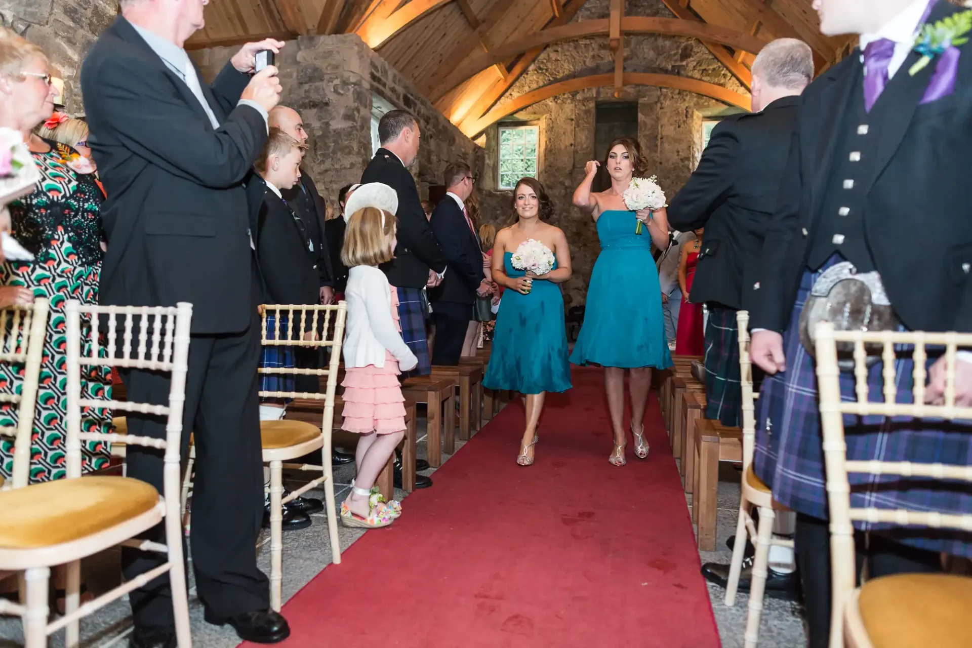 Two women in teal dresses smiling and walking down a red carpet in a stone chapel, surrounded by guests during a wedding ceremony.