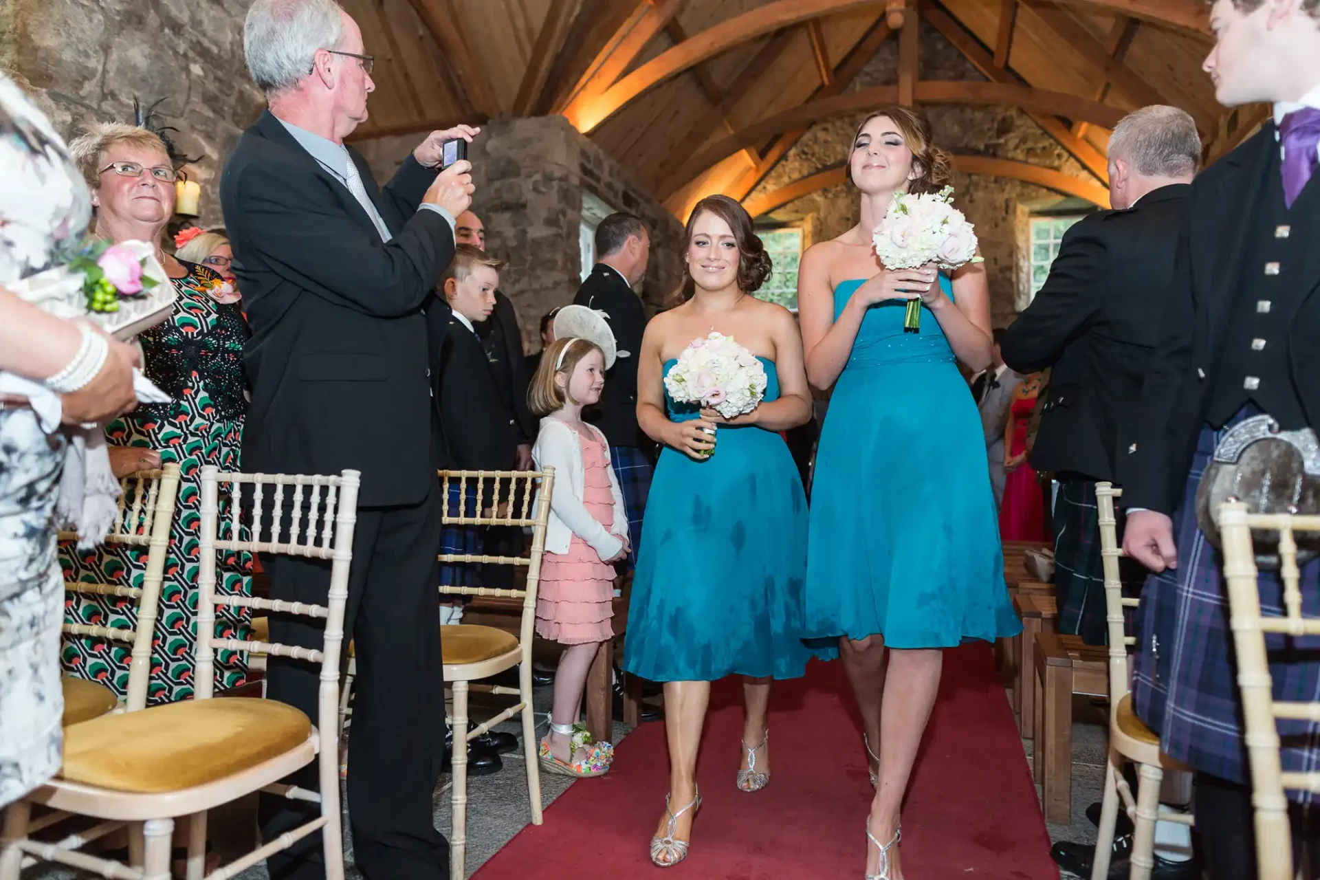 Two bridesmaids in teal dresses walk down the aisle at a wedding, holding bouquets, with guests watching and a man taking photos.