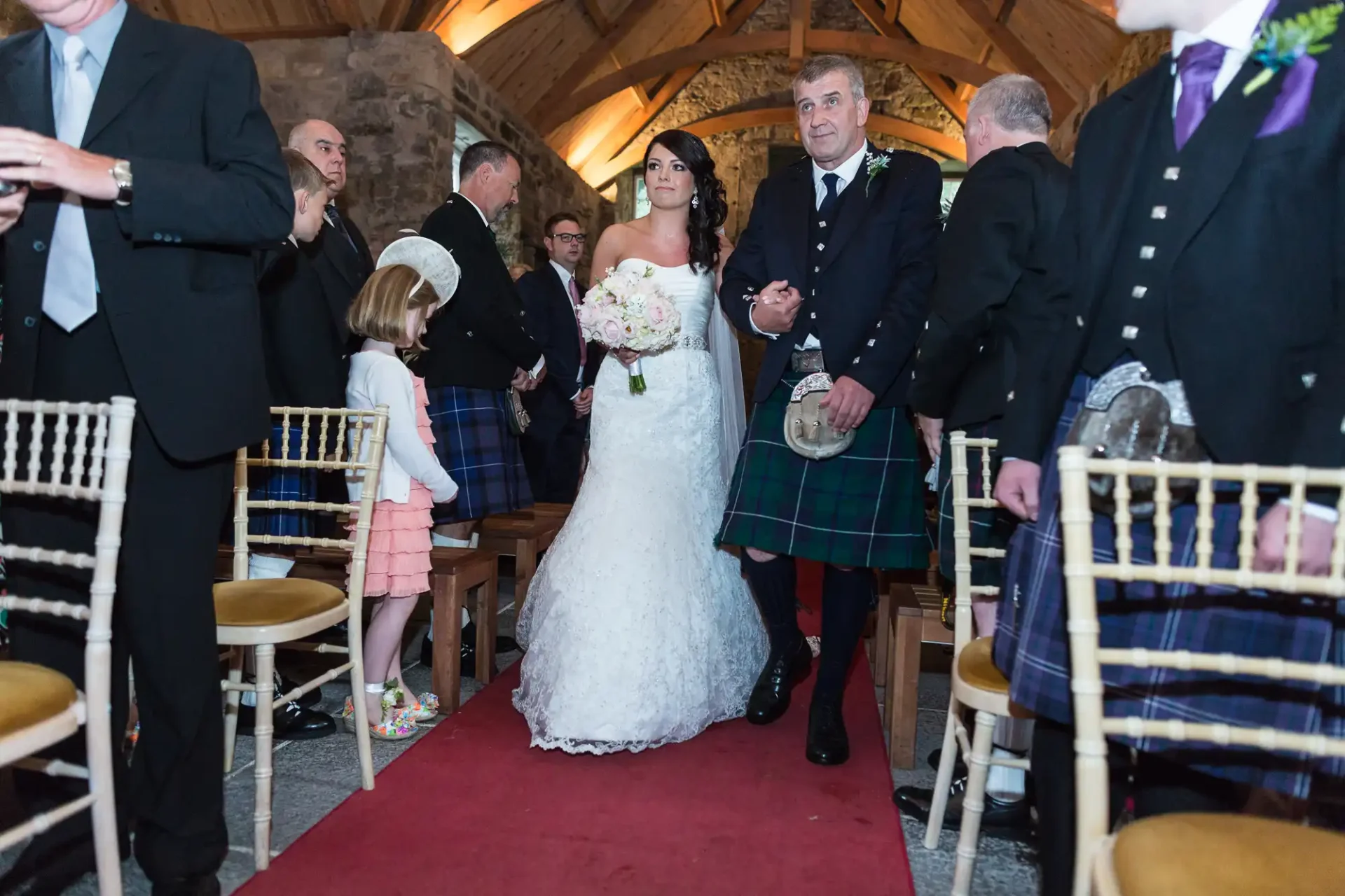 A bride in a white dress and her father wearing a kilt walk down the aisle in a stone chapel filled with guests.