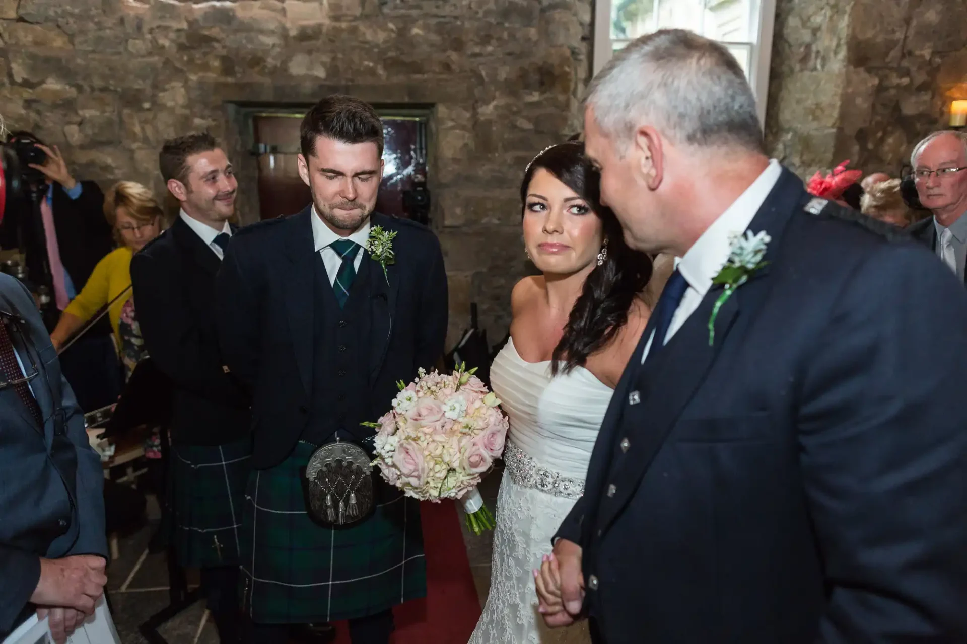 A bride and groom in a stone-walled room at a wedding ceremony, the groom in a tartan kilt and the bride holding a floral bouquet, facing a man on the right.