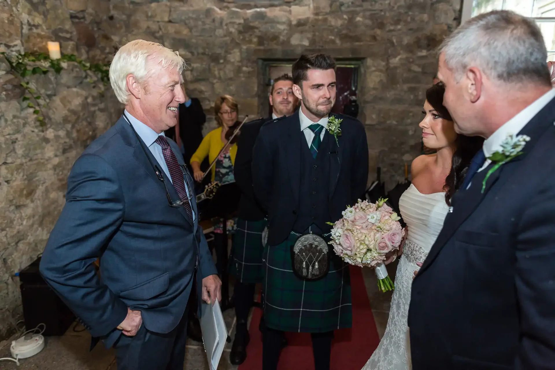 A bride and groom stand during their wedding ceremony while an older man in a suit laughs with them, another guest watches, and a man in a kilt holds the bride's bouquet.