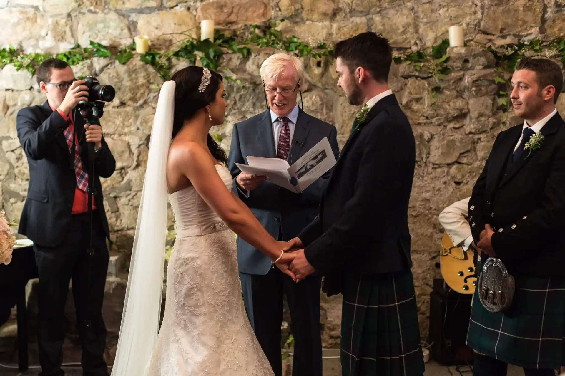 A bride and groom holding hands during their wedding ceremony, with an officiant reading vows and a photographer capturing the moment. guests in kilts are visible.