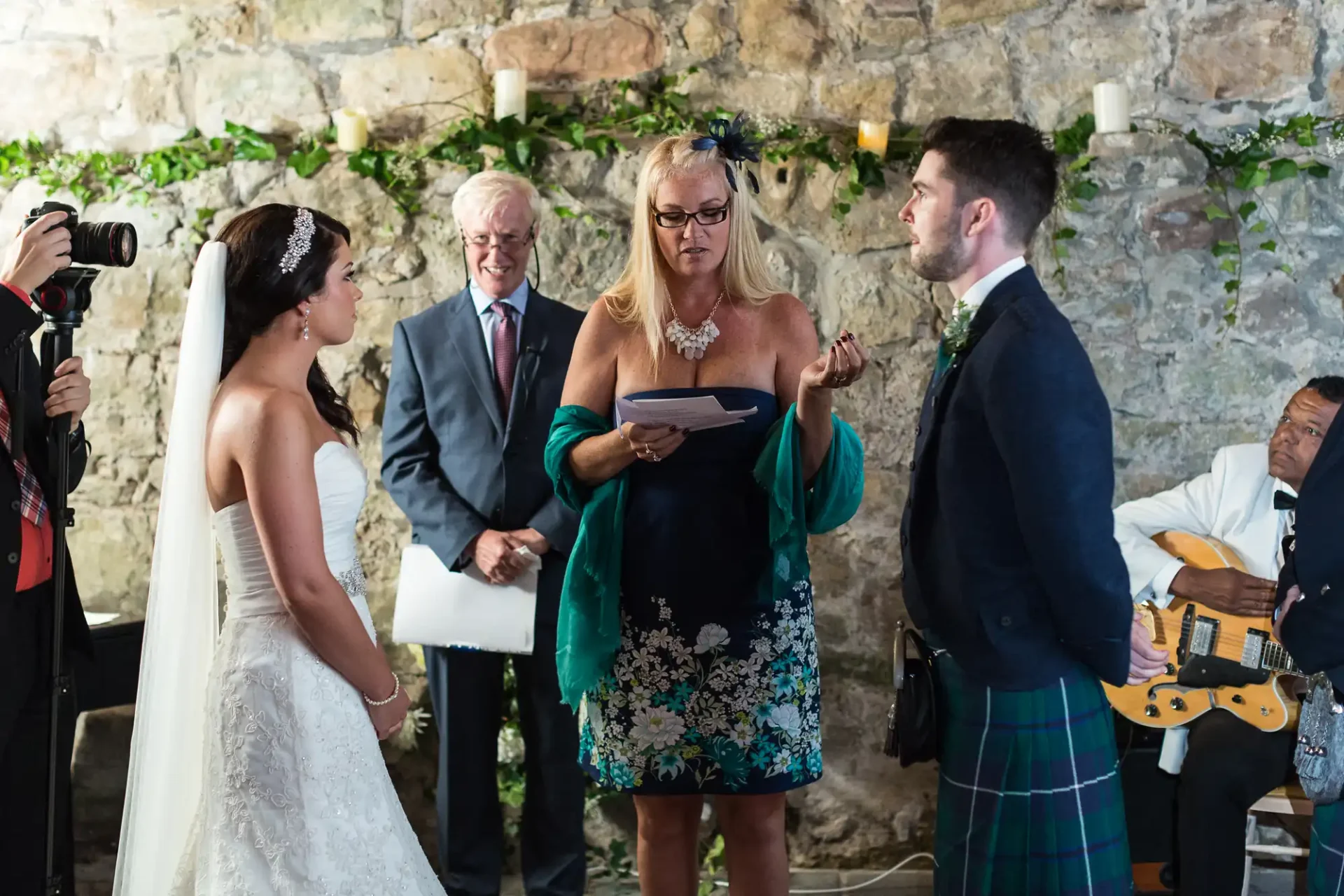 A bride and groom listen to a woman reading from a paper during a wedding ceremony, with an officiant and photographer nearby, in a rustic stone venue.