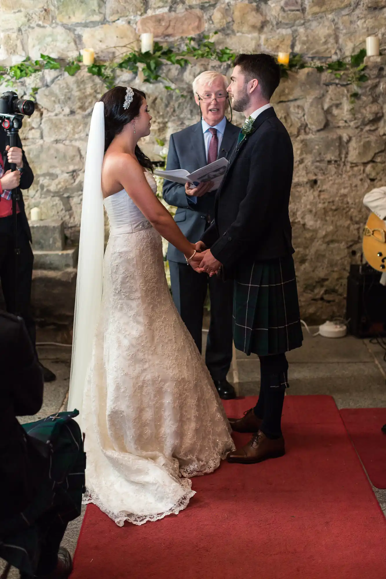 Bride in a lace gown and groom in a kilt holding hands during a wedding ceremony, with an officiant speaking and a photographer capturing the moment.