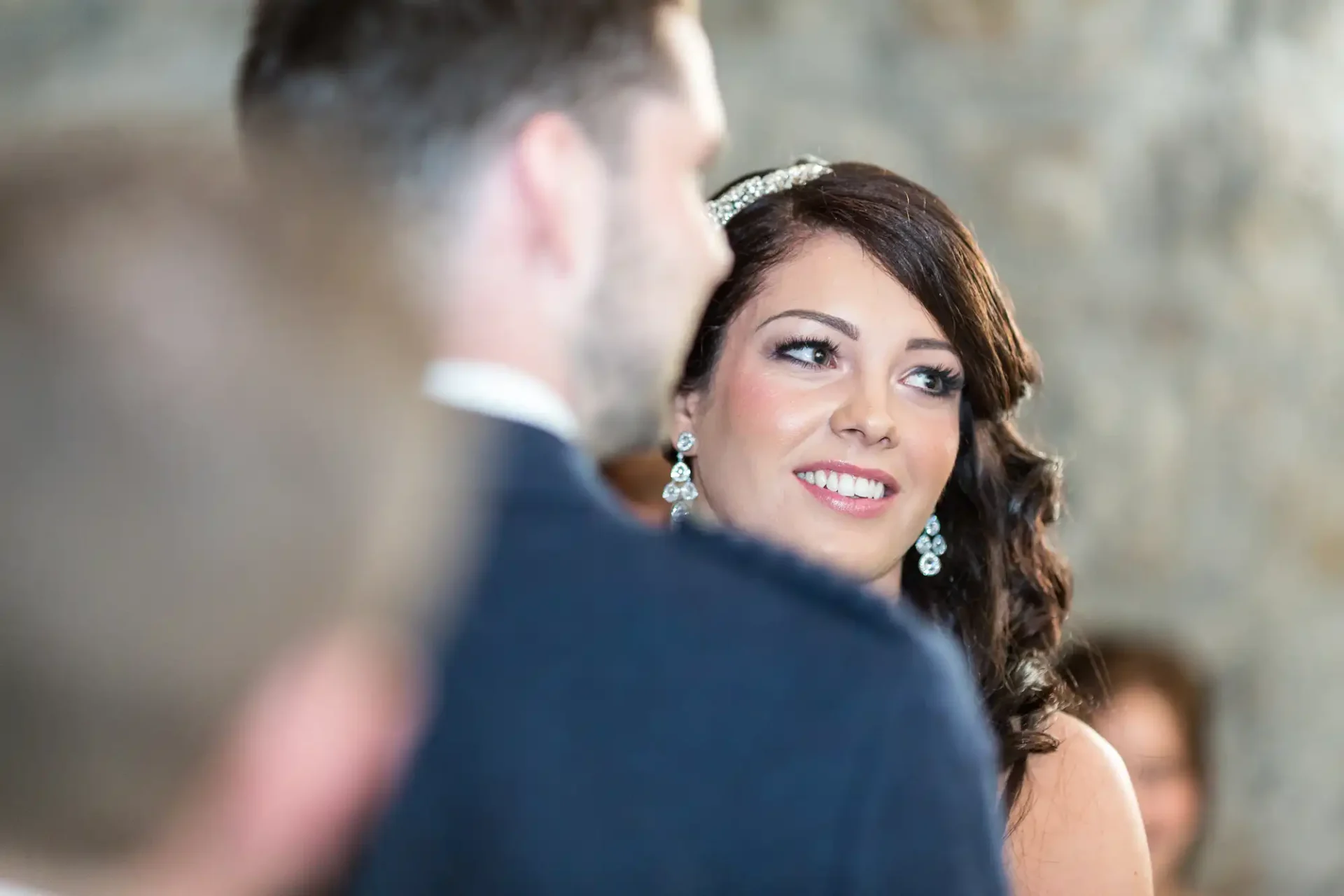 A bride in a tiara smiles at a groom, who is seen from behind, during a wedding ceremony.