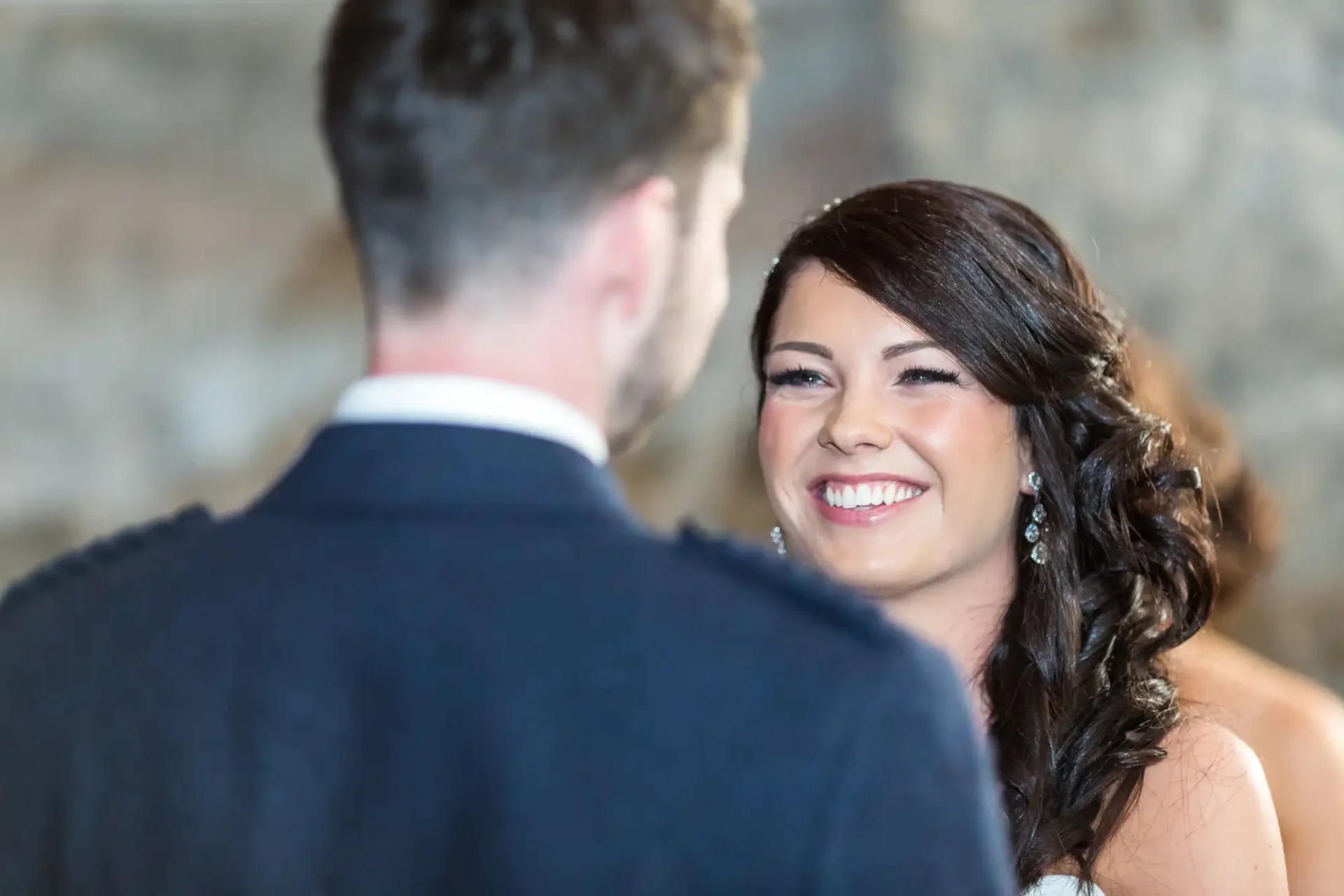 A smiling bride looking at the groom, seen from over his shoulder, in a softly lit indoor setting.