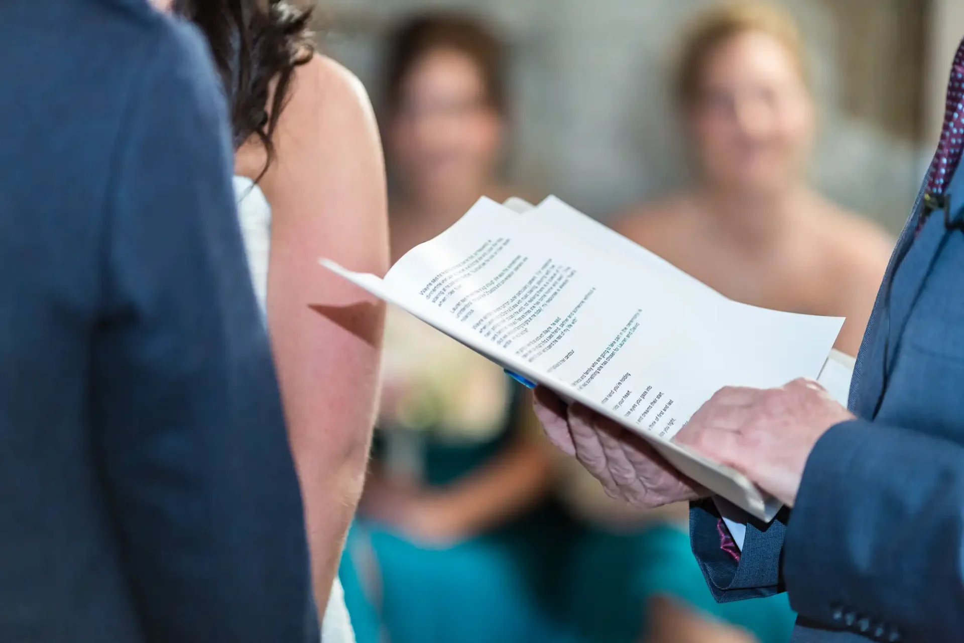 A close-up of a wedding vow sheet held by a groom in front of a bride, with bridesmaids slightly blurred in the background.