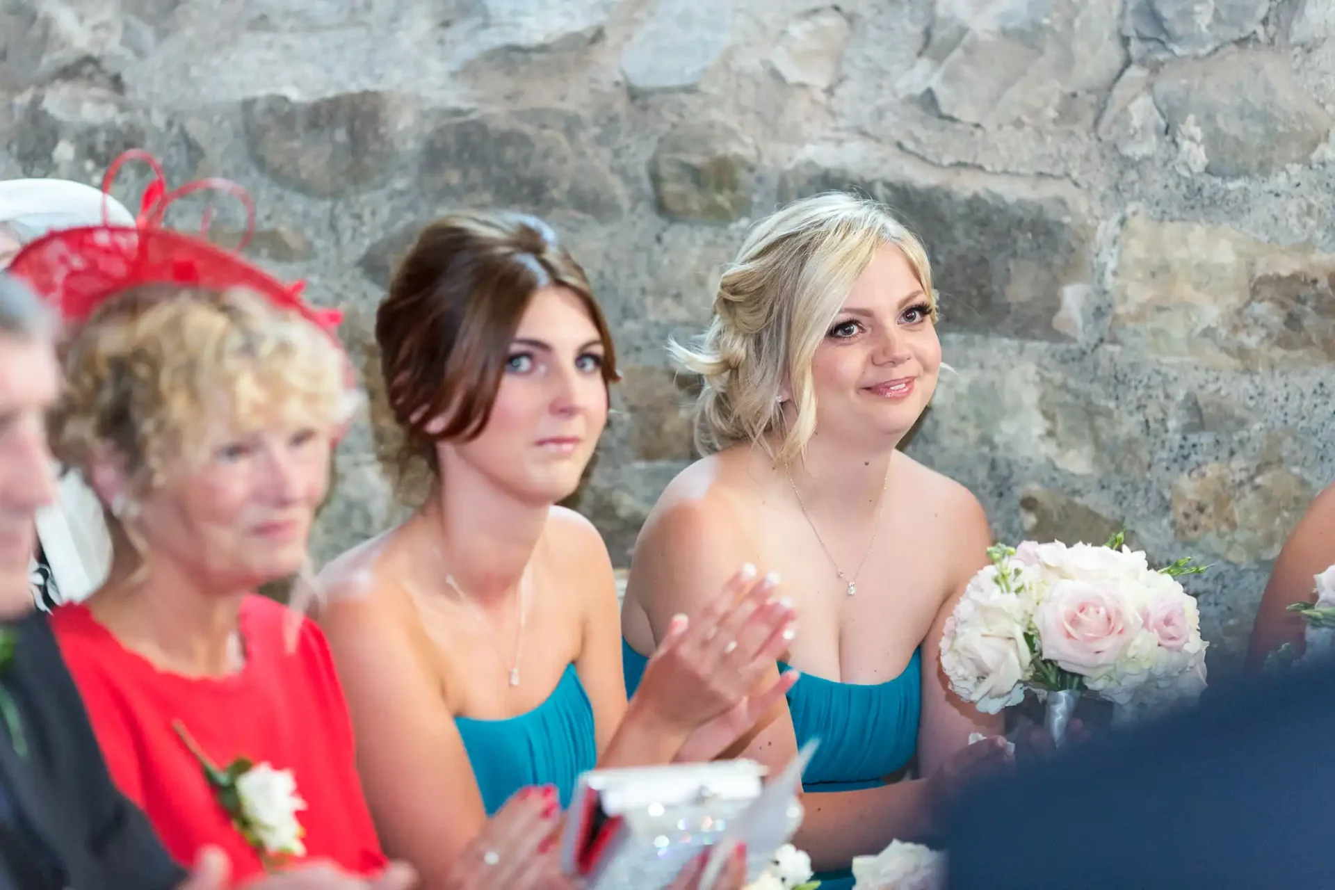 Three women at a wedding ceremony, wearing elegant dresses and sitting by a table adorned with flowers, clapping and looking attentively towards the front.