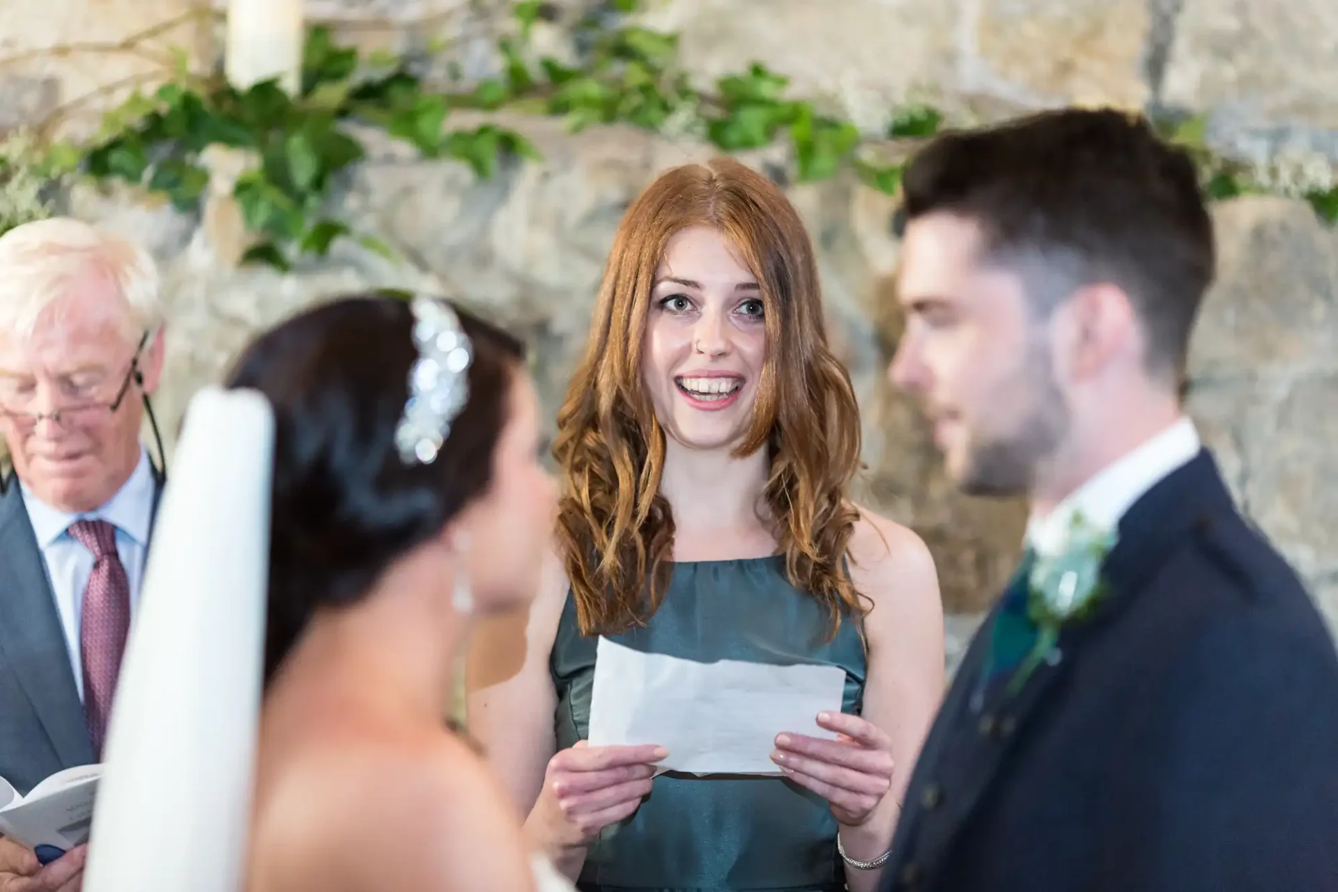 Woman in green dress reading a speech at a wedding, smiling at the bride and groom, with an older man in the background.
