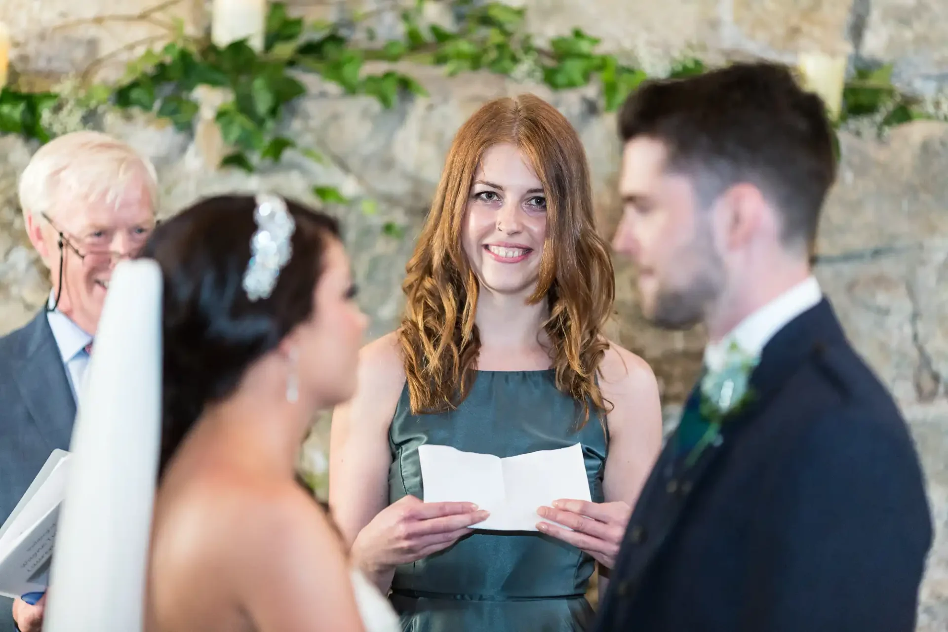 A woman in a green dress gives a speech at a wedding, with the bride and groom listening attentively in the foreground.