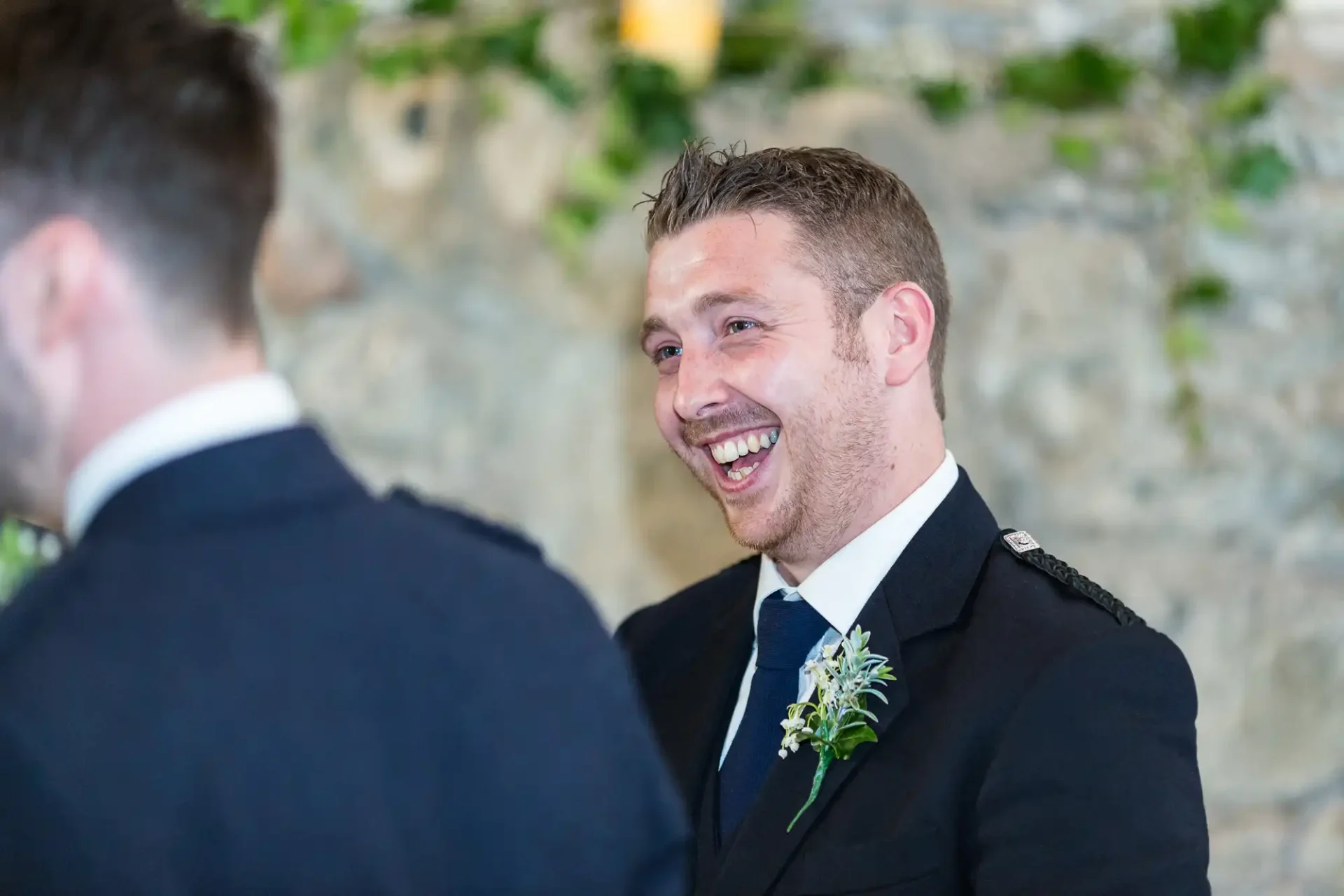 Man in a suit with a boutonniere laughing joyfully at a wedding event, facing another man with his back turned to the camera.