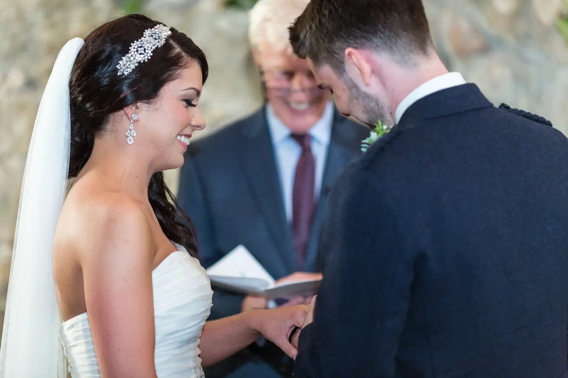 A bride and groom exchanging vows, smiling at each other, with an officiant looking on in the background.