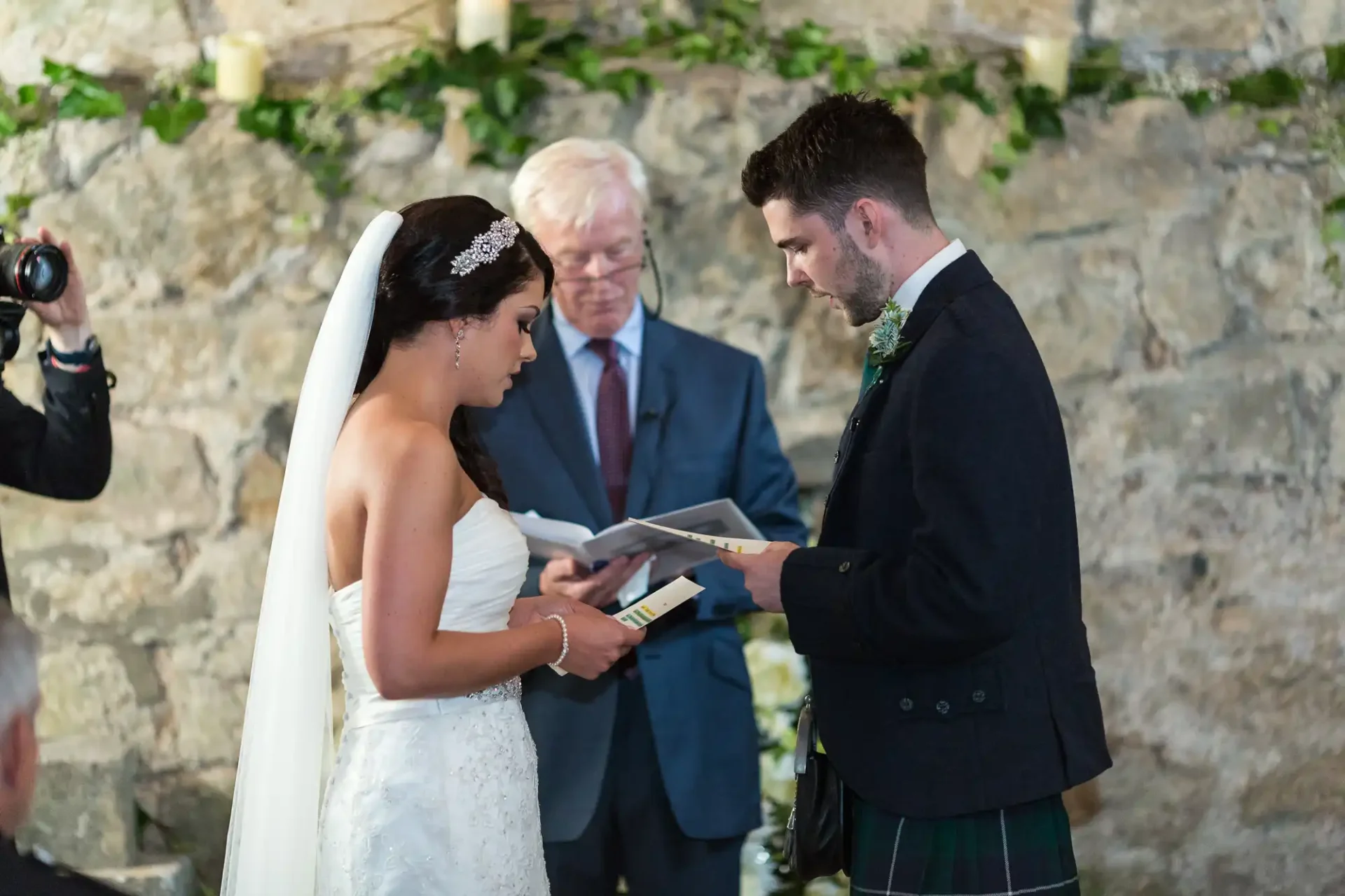 A bride and groom exchange vows, with the groom in a kilt, while an officiant watches and a photographer captures the moment in a rustic venue.