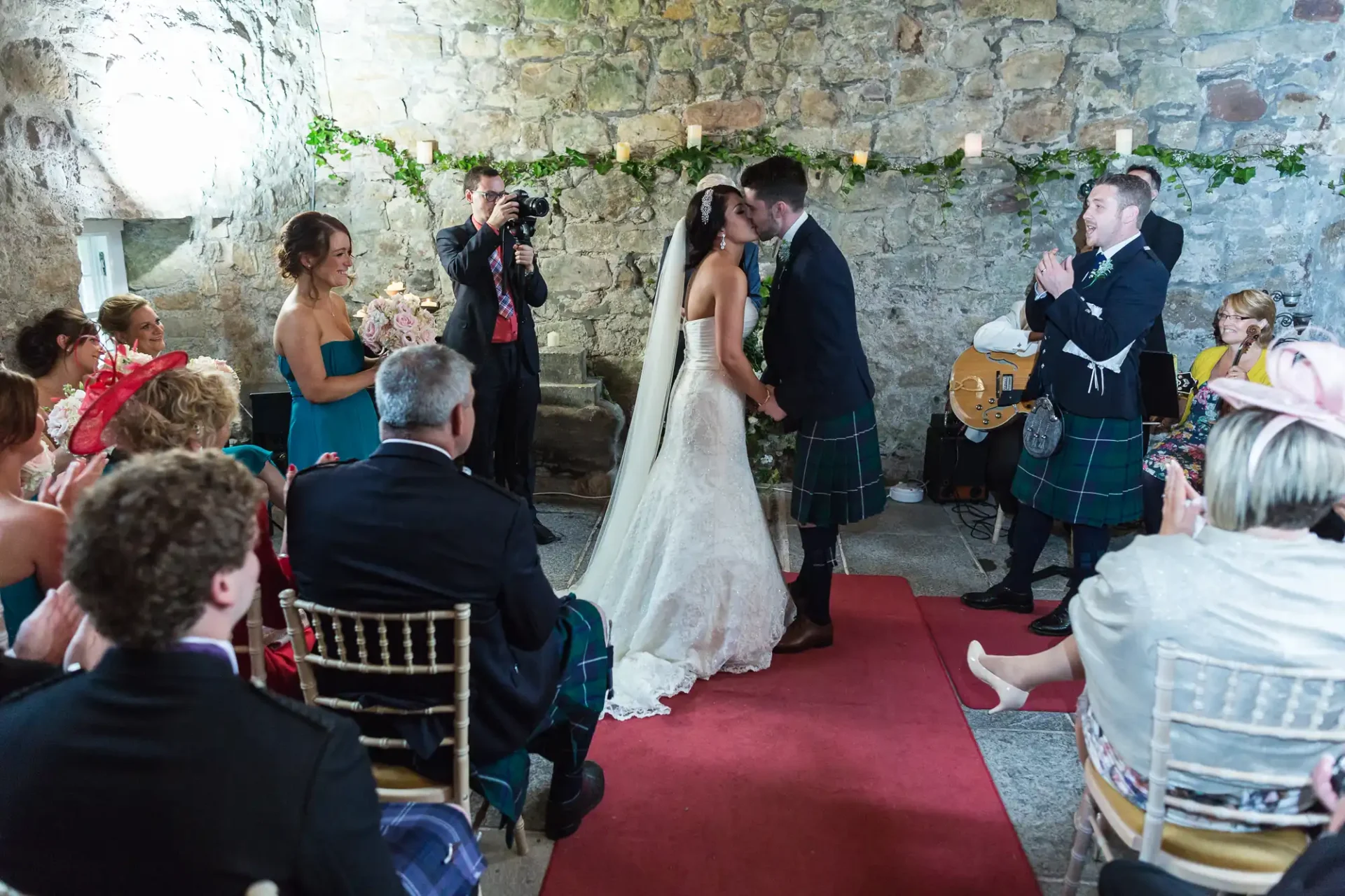A bride and groom kissing at their wedding ceremony inside a rustic stone building, with guests and a photographer capturing the moment.