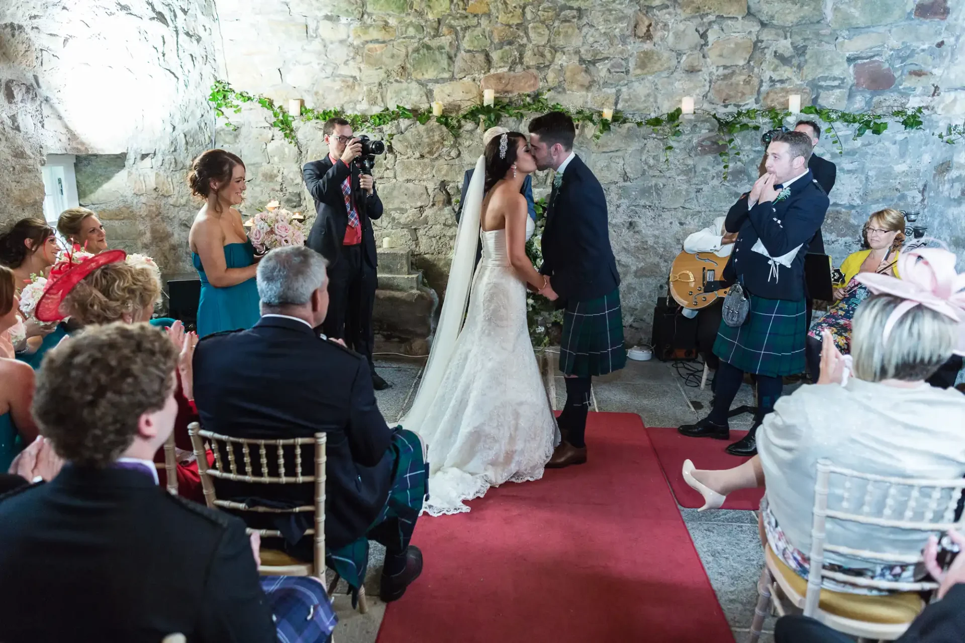 A bride and groom kiss during their wedding ceremony in a rustic stone hall, surrounded by guests and musicians wearing kilts.