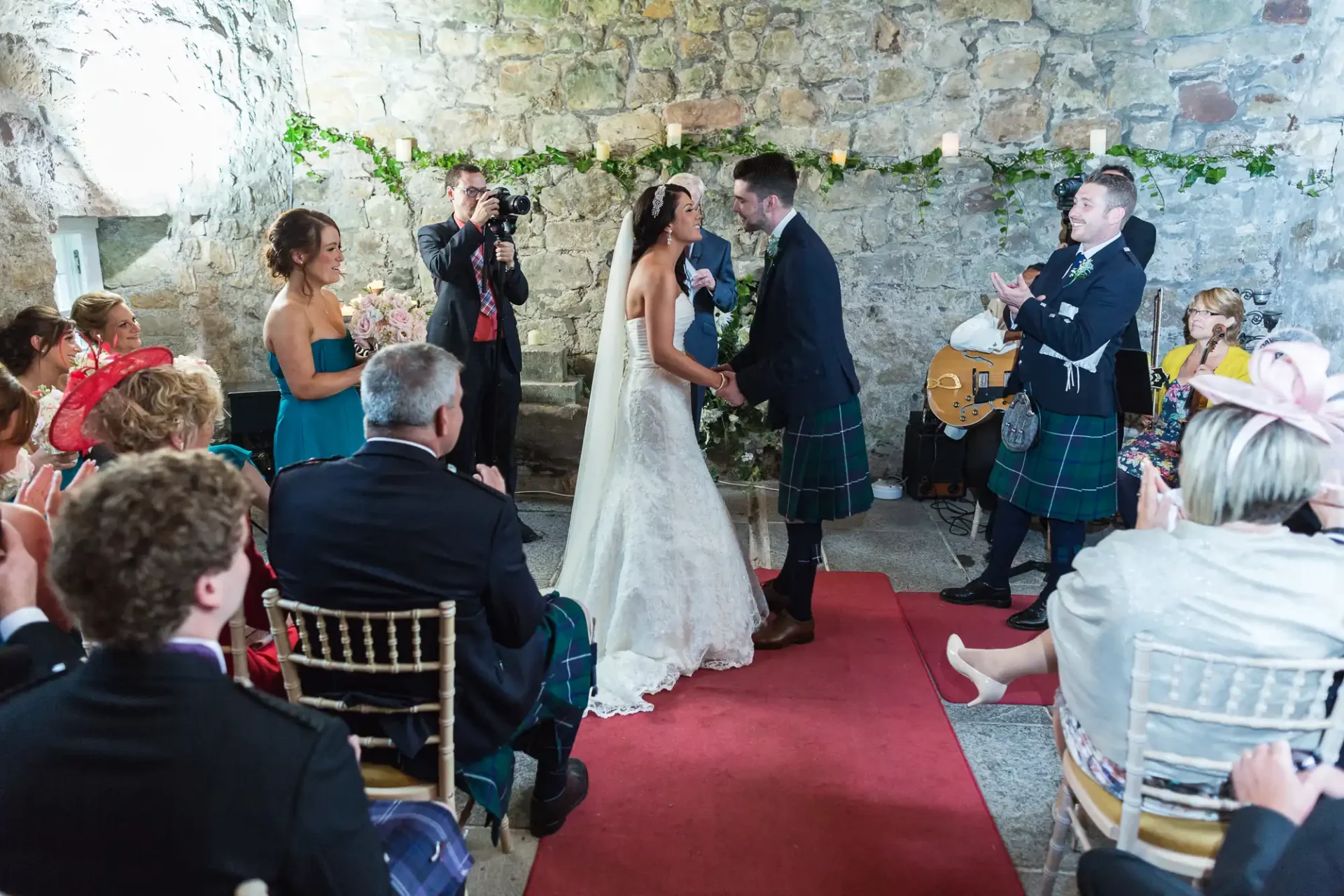 A bride and groom exchange vows at an intimate indoor wedding ceremony, with guests in formal attire and a musician playing the bagpipes.