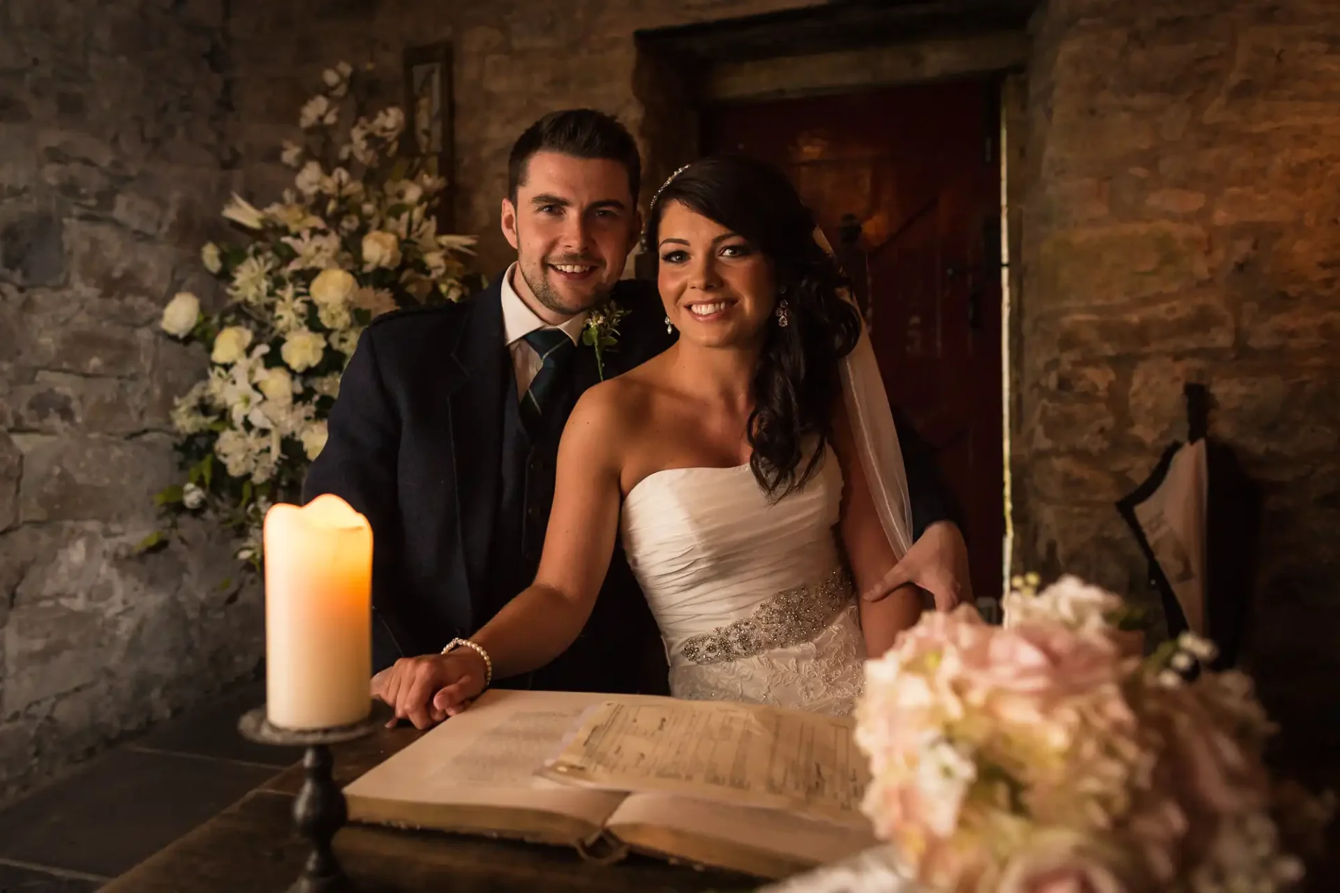 A newlywed couple signing a marriage register in a candlelit room, surrounded by flowers.