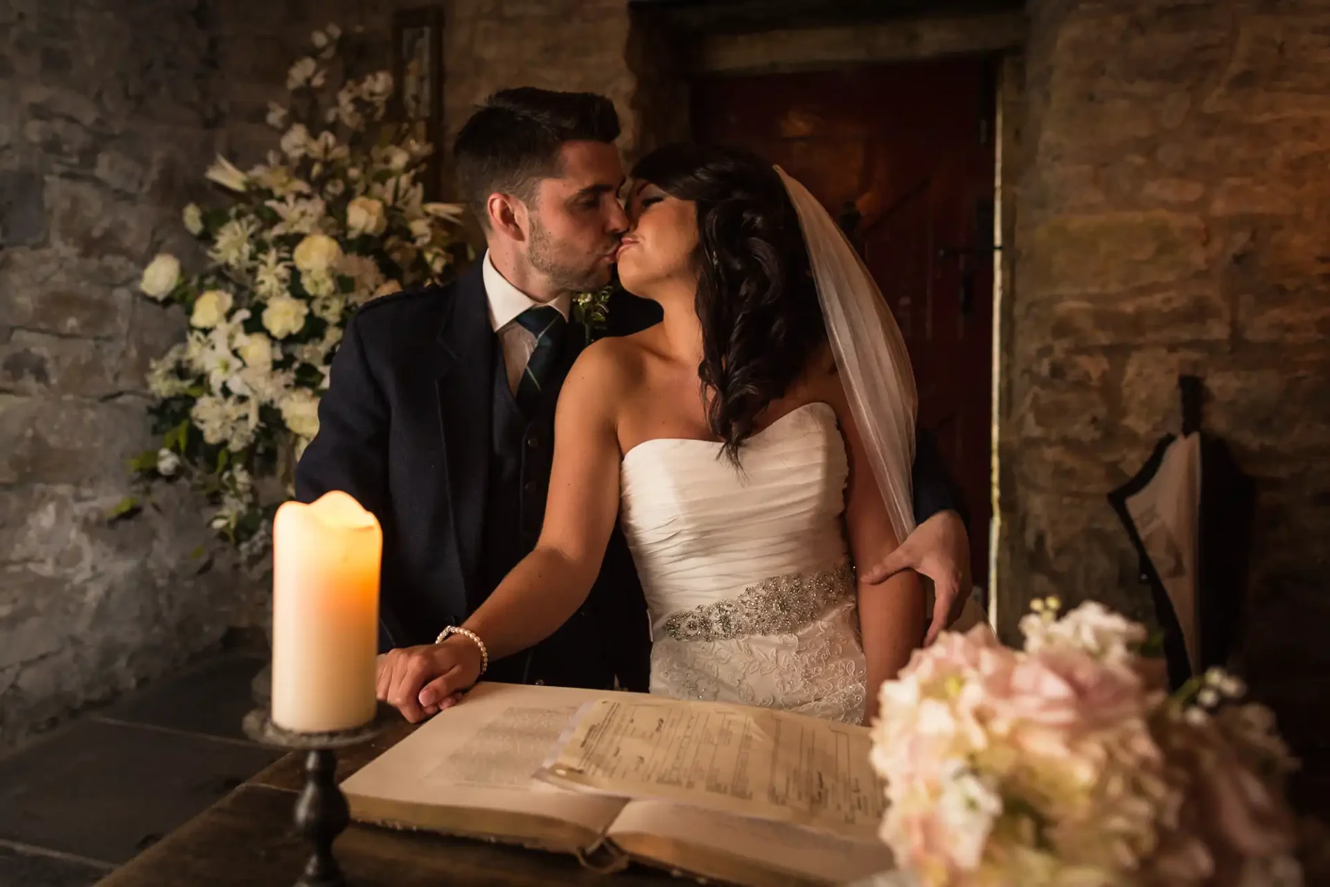 Bride and groom kissing at the signing table, surrounded by candles and flowers in a dimly lit rustic venue.