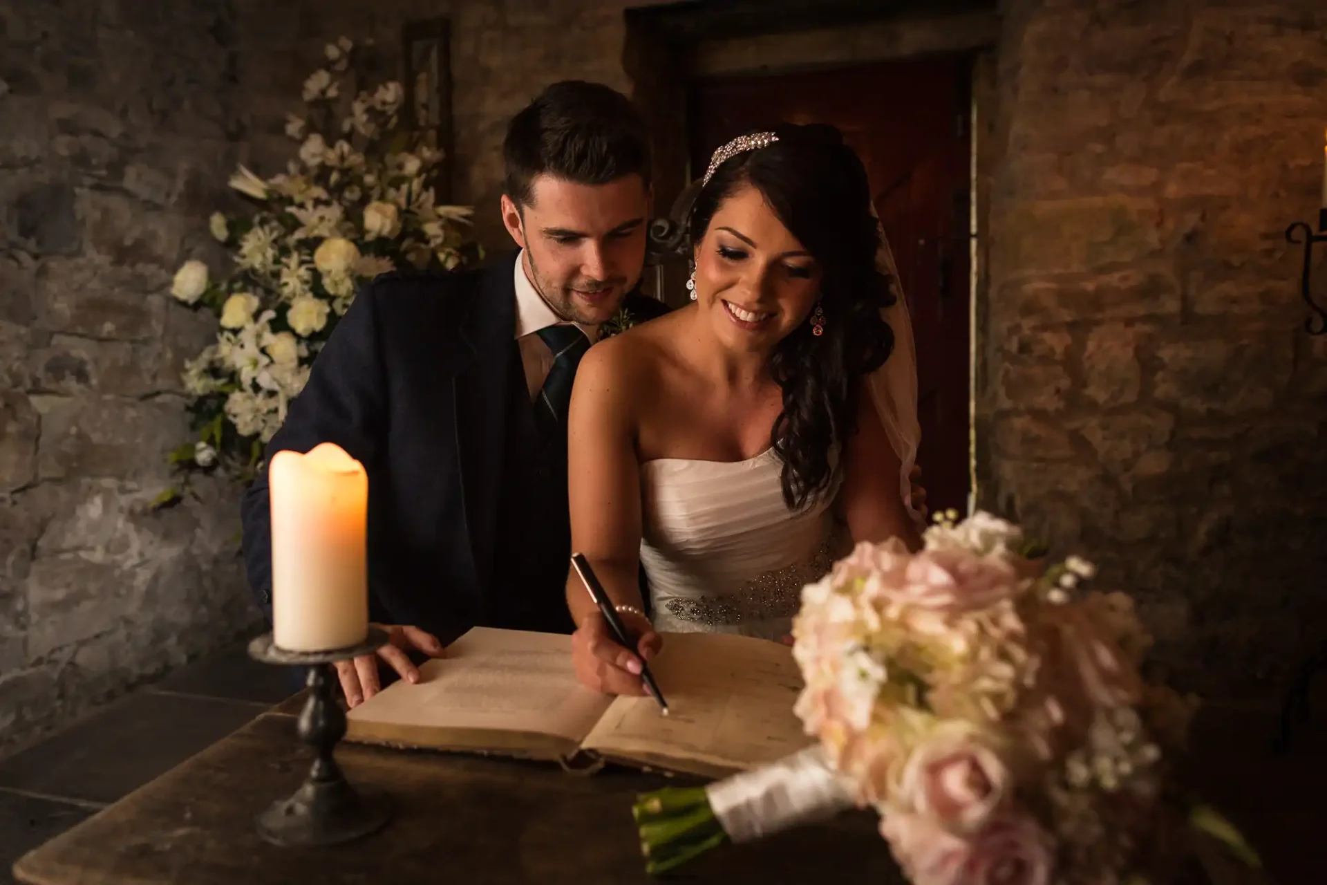 Bride in a white dress and groom in a suit signing a marriage register in a warmly lit, stone-walled room with candles and floral decorations.