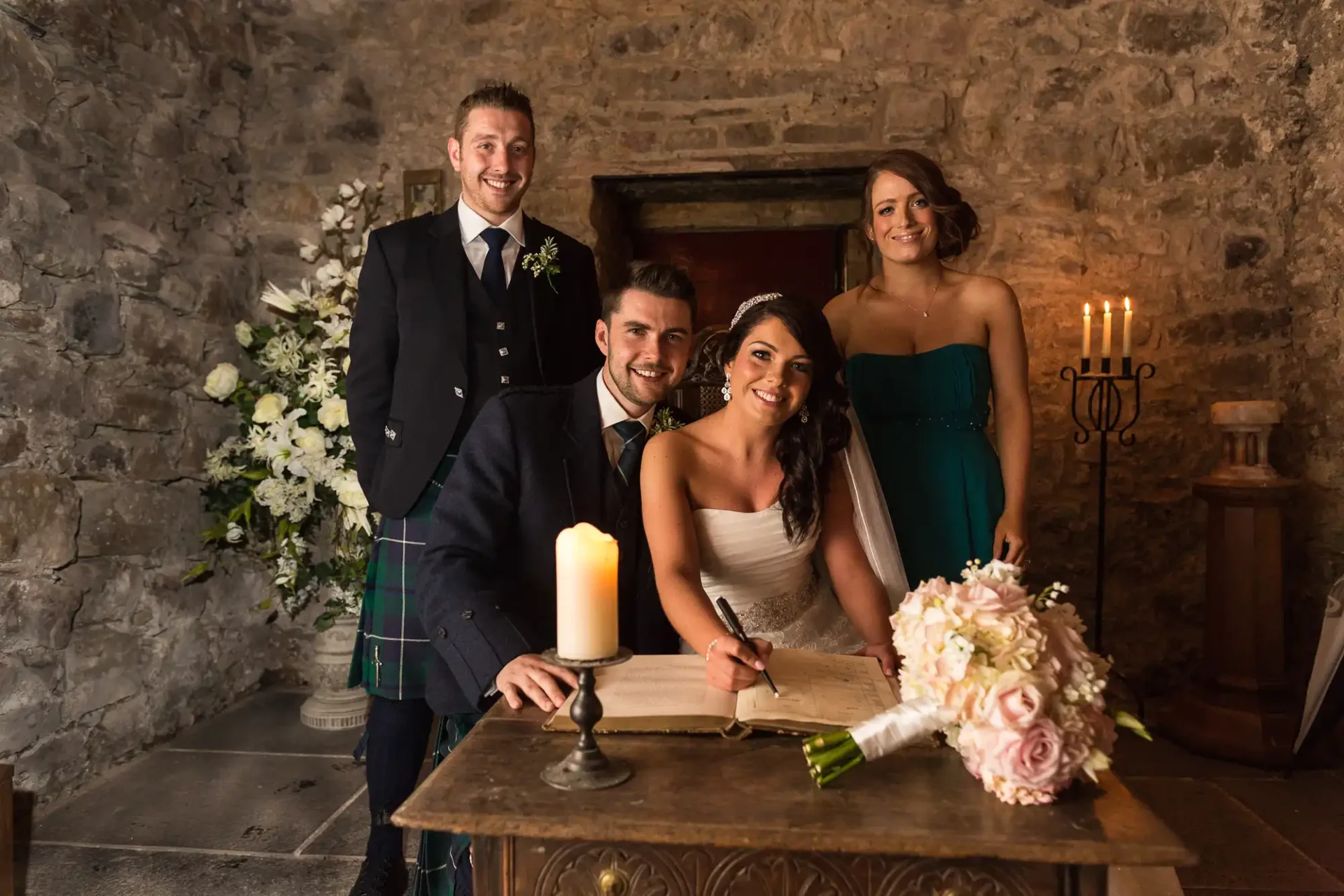 A newlywed couple signs a marriage register flanked by a best man and a bridesmaid in a candle-lit stone room.