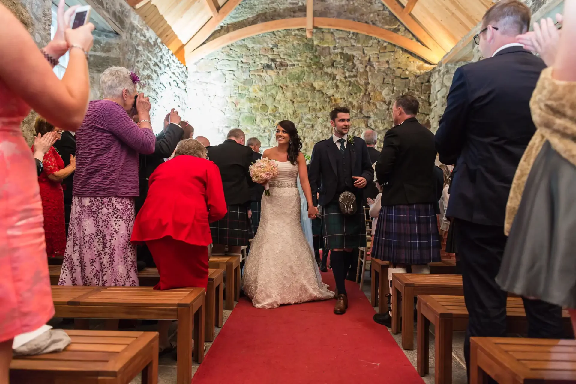 A bride and groom happily walk down the aisle of a stone-walled room, surrounded by guests, some wearing traditional scottish kilts.