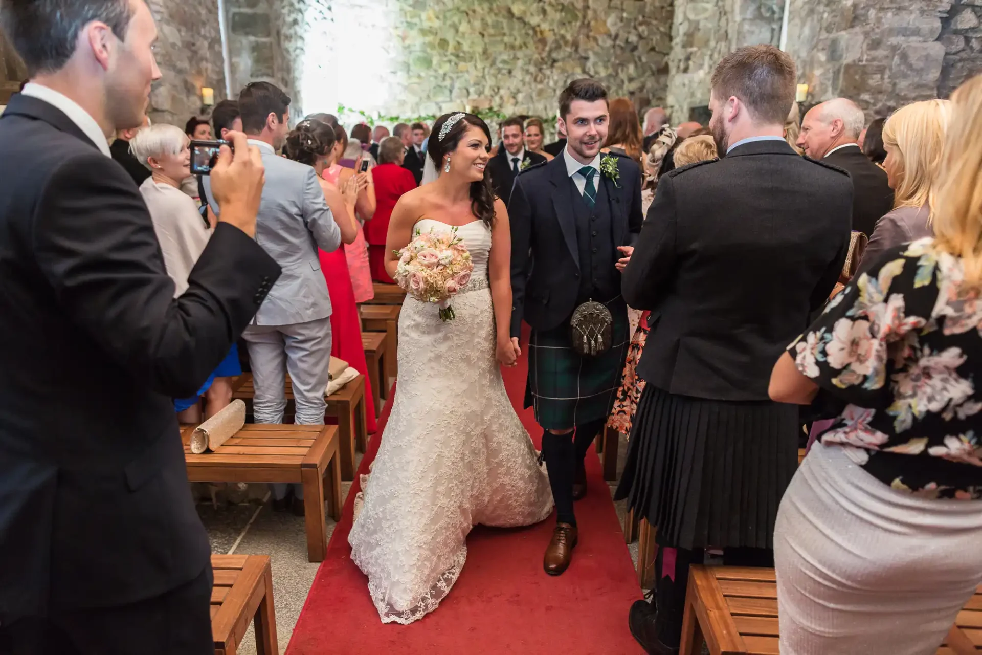 A bride and groom smiling and walking down the aisle, surrounded by guests, inside a stone-walled church. the groom wears a kilt.