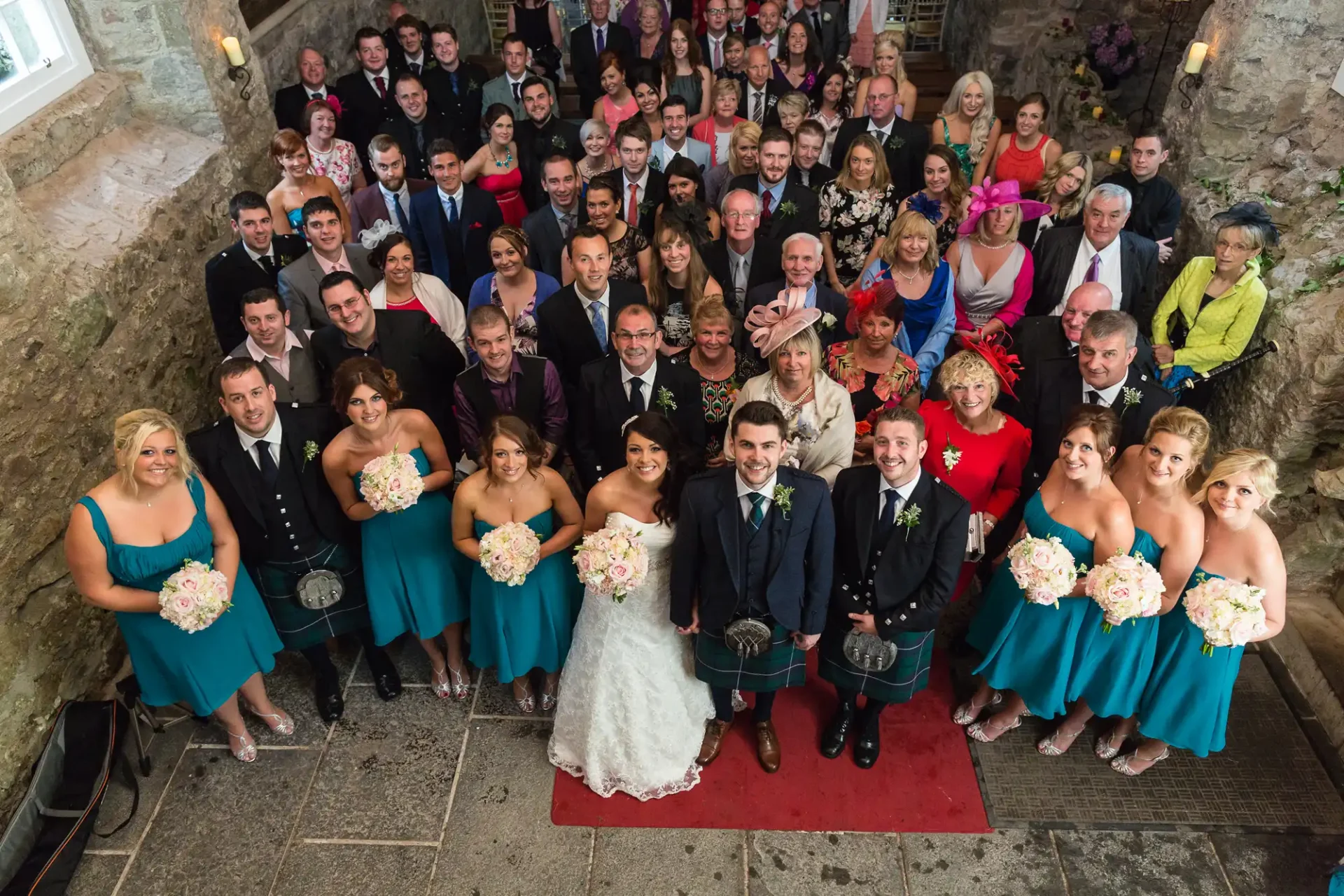 A large wedding group posing on steps, with a couple in front; bridesmaids wear teal dresses, men in kilts.