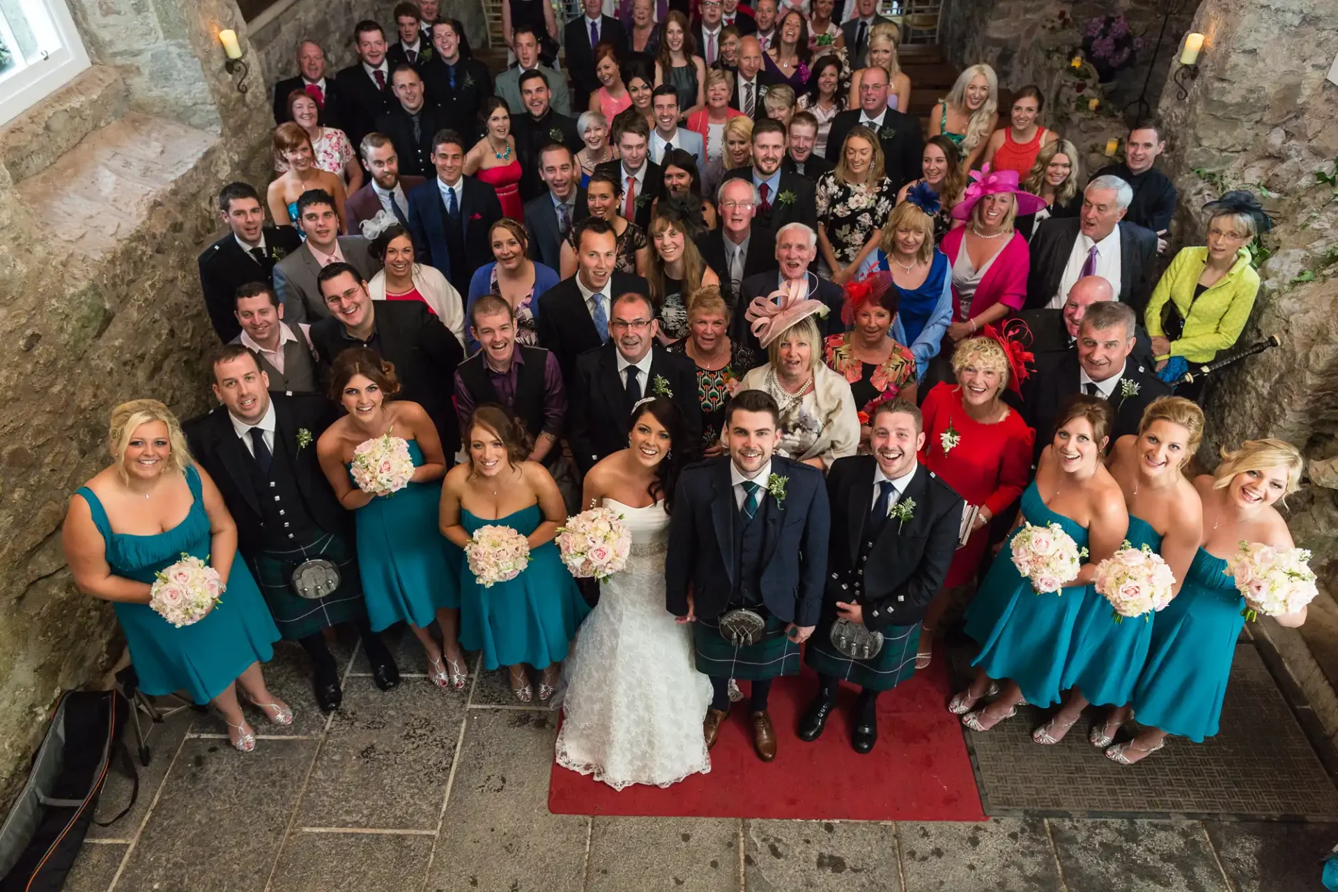 A large group of people posing for a wedding photo on a stone staircase, with several individuals in vibrant teal dresses and dark suits, centered by a bride and groom.