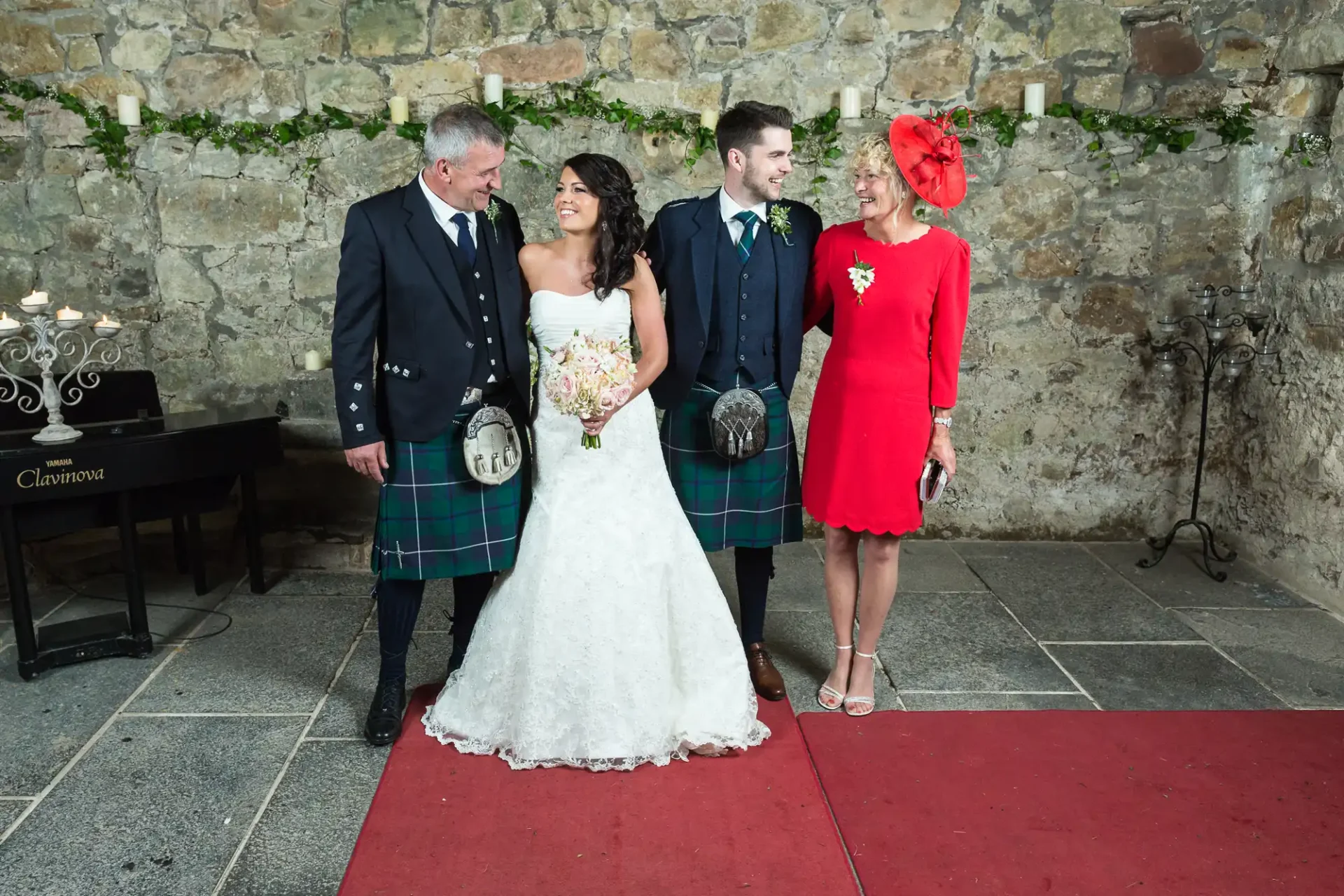 A bride and groom stand with two guests at their wedding in a stone-walled hall, the men in traditional kilts and the women in dresses.