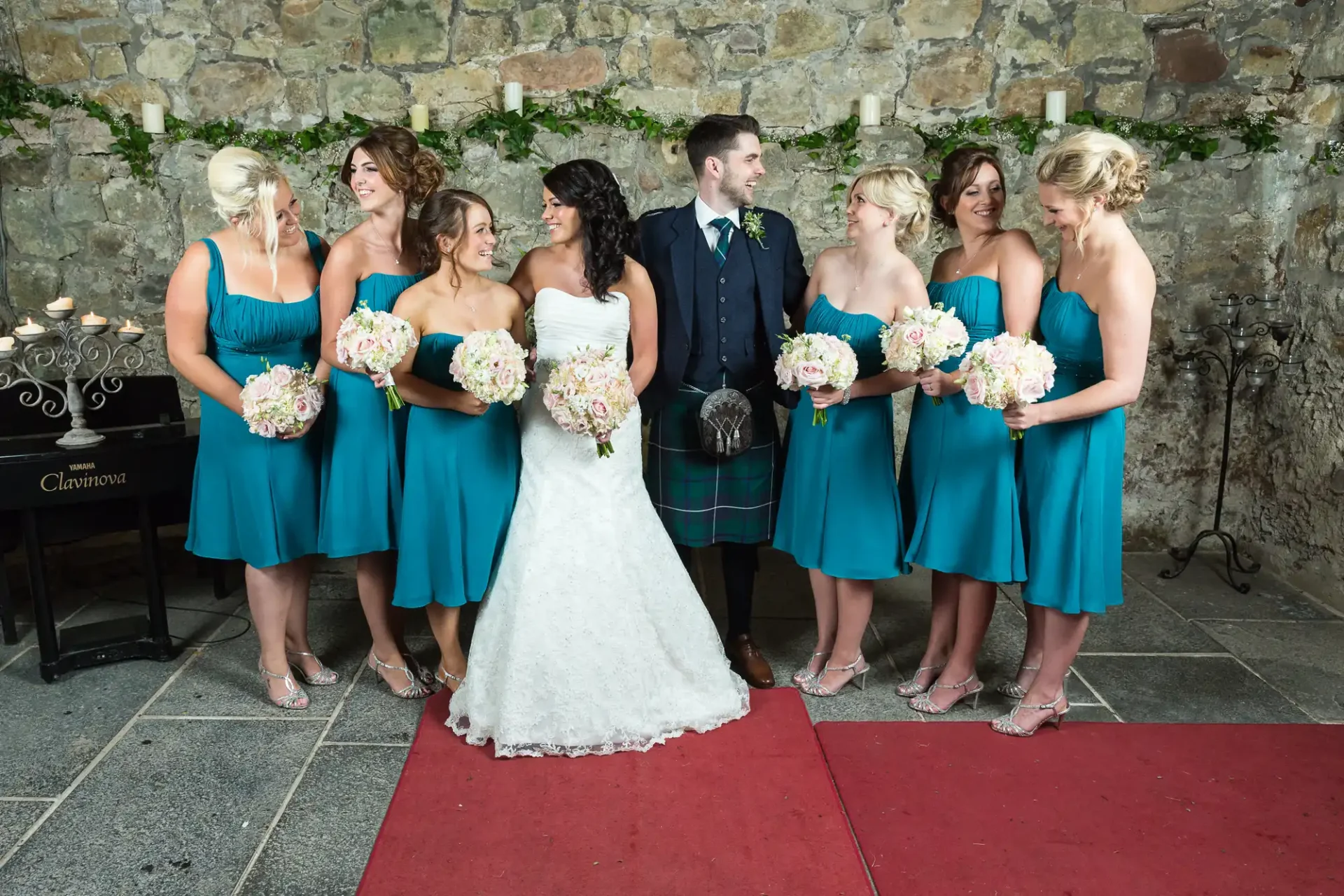 A bride and groom posing with bridesmaids dressed in teal, all holding bouquets, in front of a stone wall.