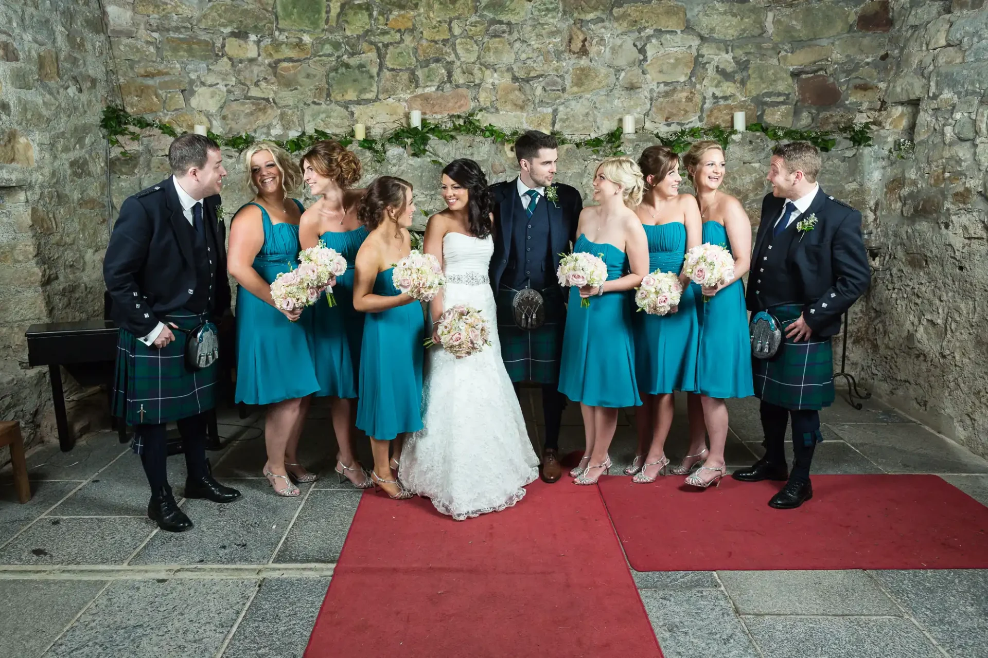 Wedding group in semi-formal attire, with men in kilts and a bride in a white dress, standing against a stone wall, smiling and interacting.