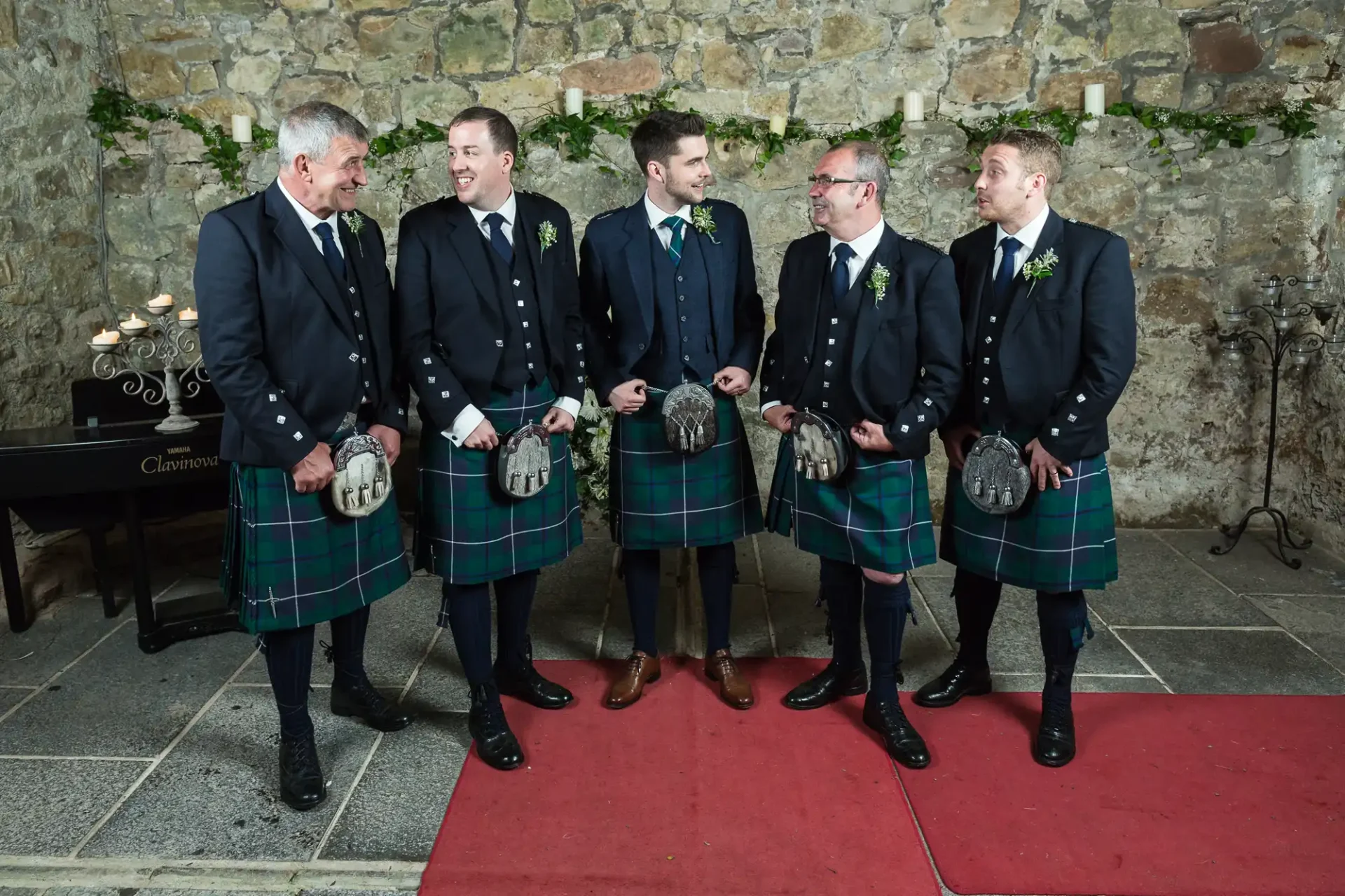 Five men in traditional scottish kilts and jackets stand on a red carpet against a stone wall, talking and smiling at a formal event.
