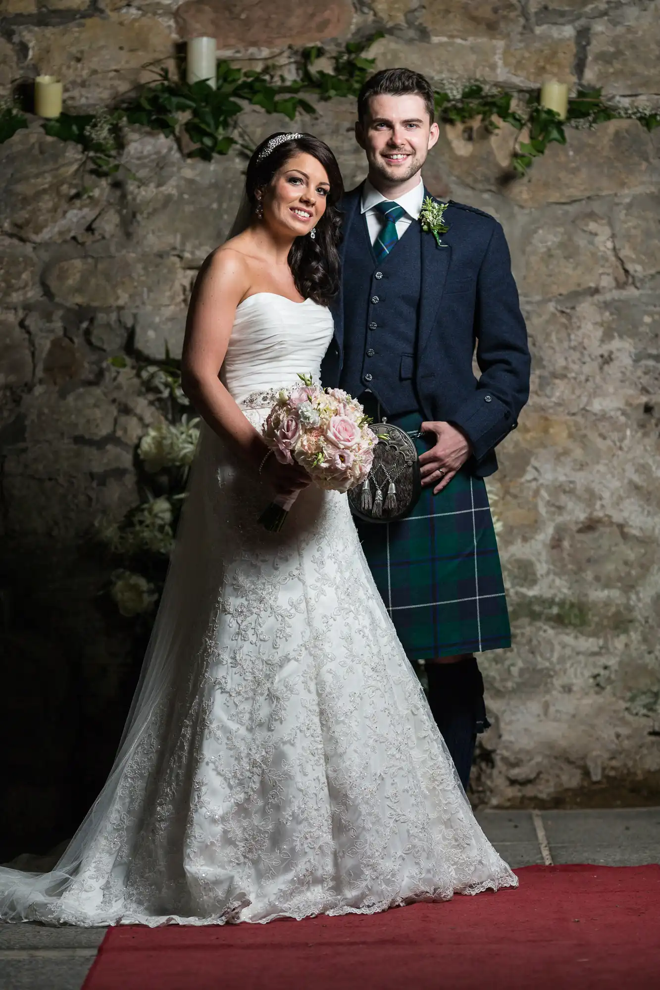 A bride and groom smiling, standing together in a traditional wedding attire; the bride in a white dress and the groom in a kilt.
