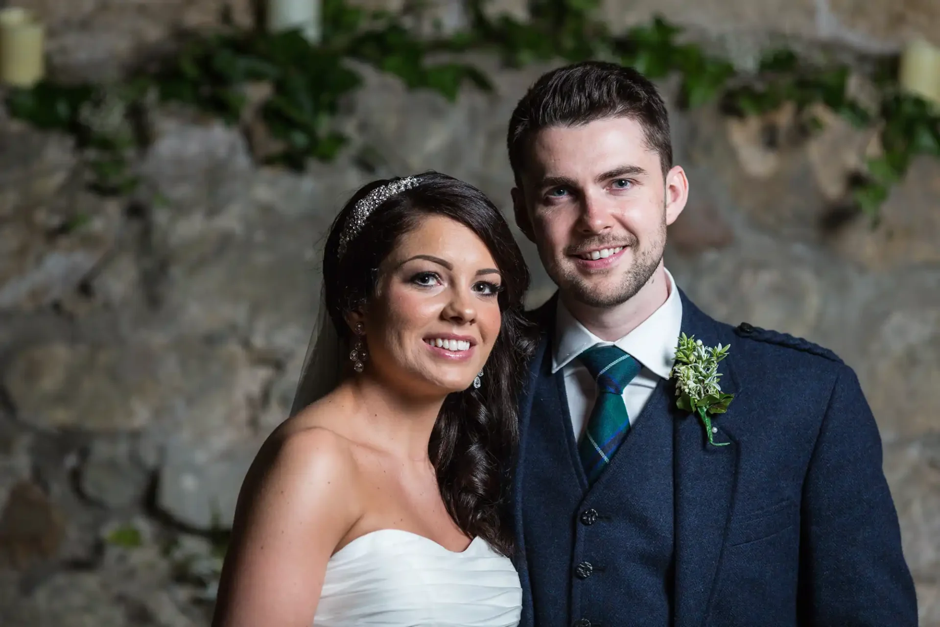 A bride and groom smiling for a photo, the bride in a white strapless dress and the groom in a dark suit with a tartan tie and green boutonniere.