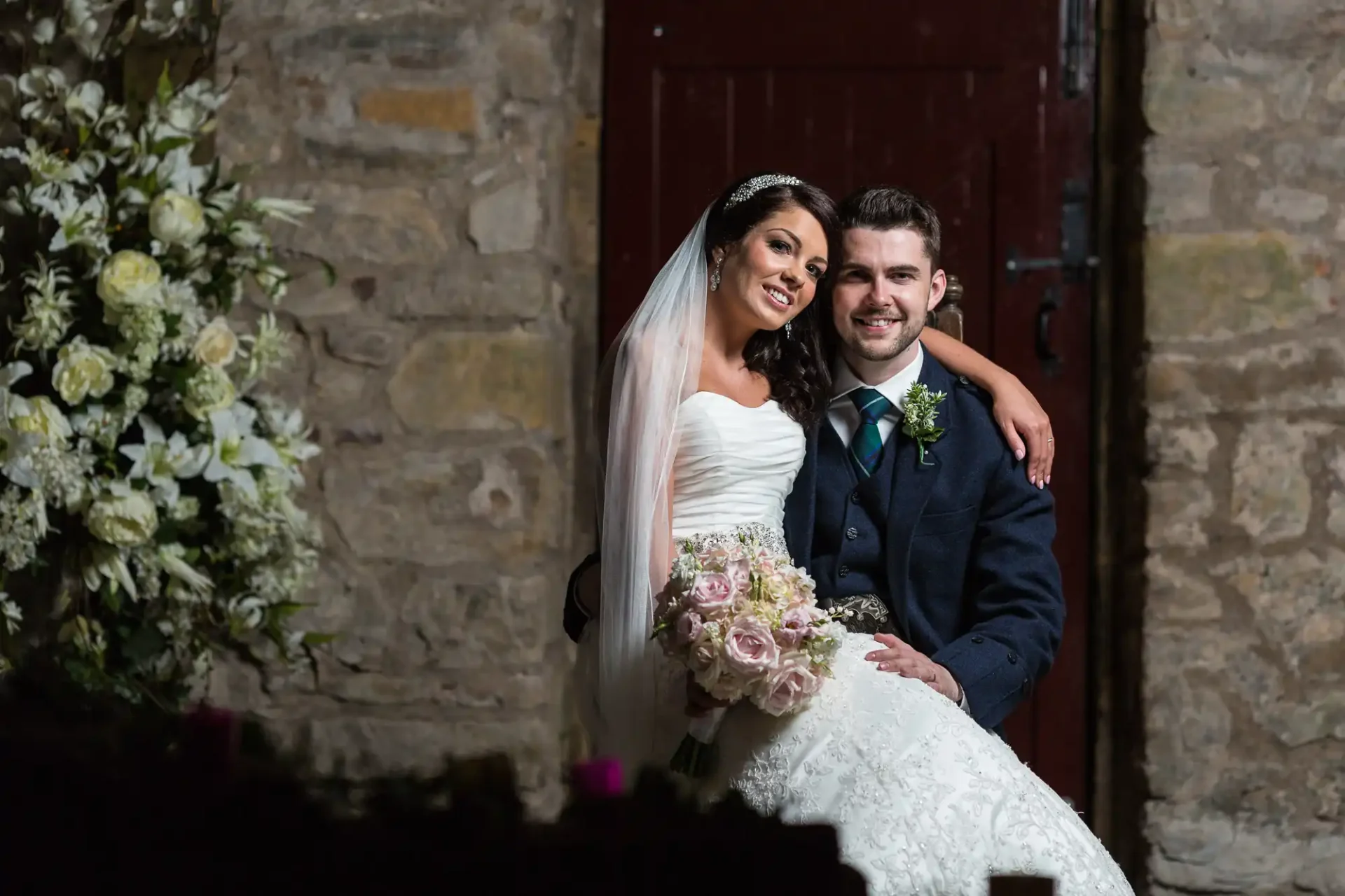 A bride and groom smiling and posing together at night, with the bride holding a bouquet, set against a stone building with a dark doorway.
