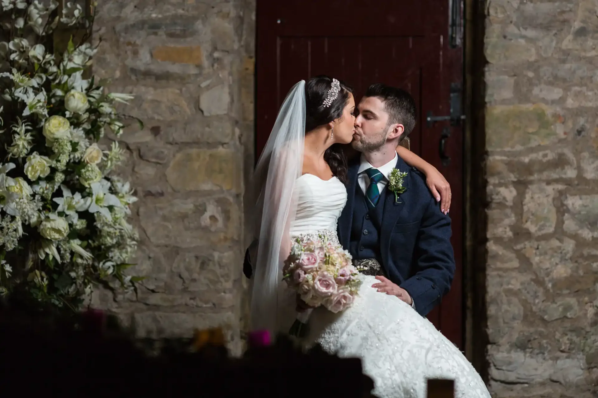 A bride and groom kiss outside a stone building, with the bride holding a bouquet of pink and white flowers.