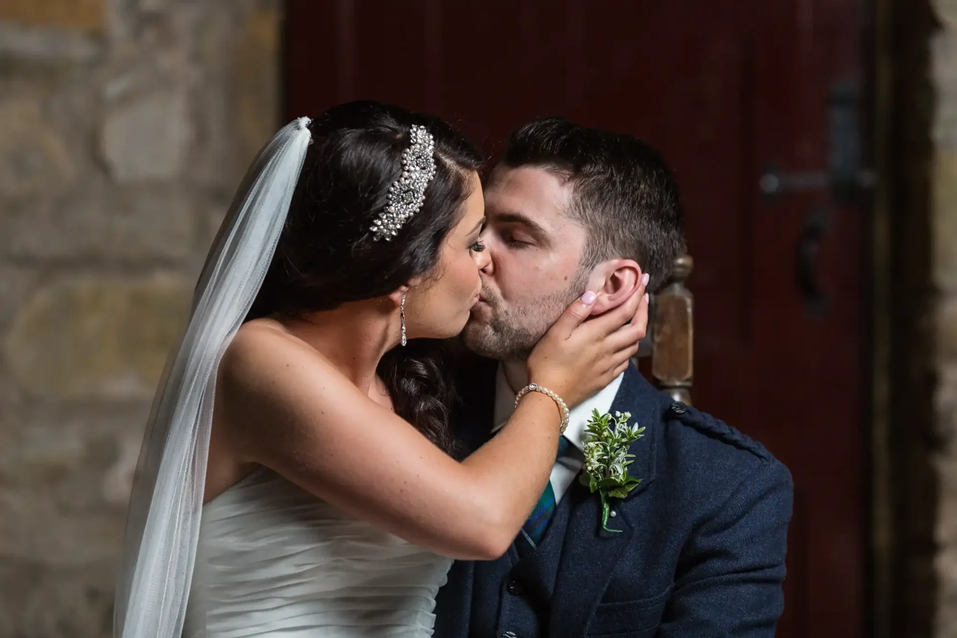 A bride in a white dress and a groom in a blue suit sharing a kiss, with a stone wall and wooden door in the background.