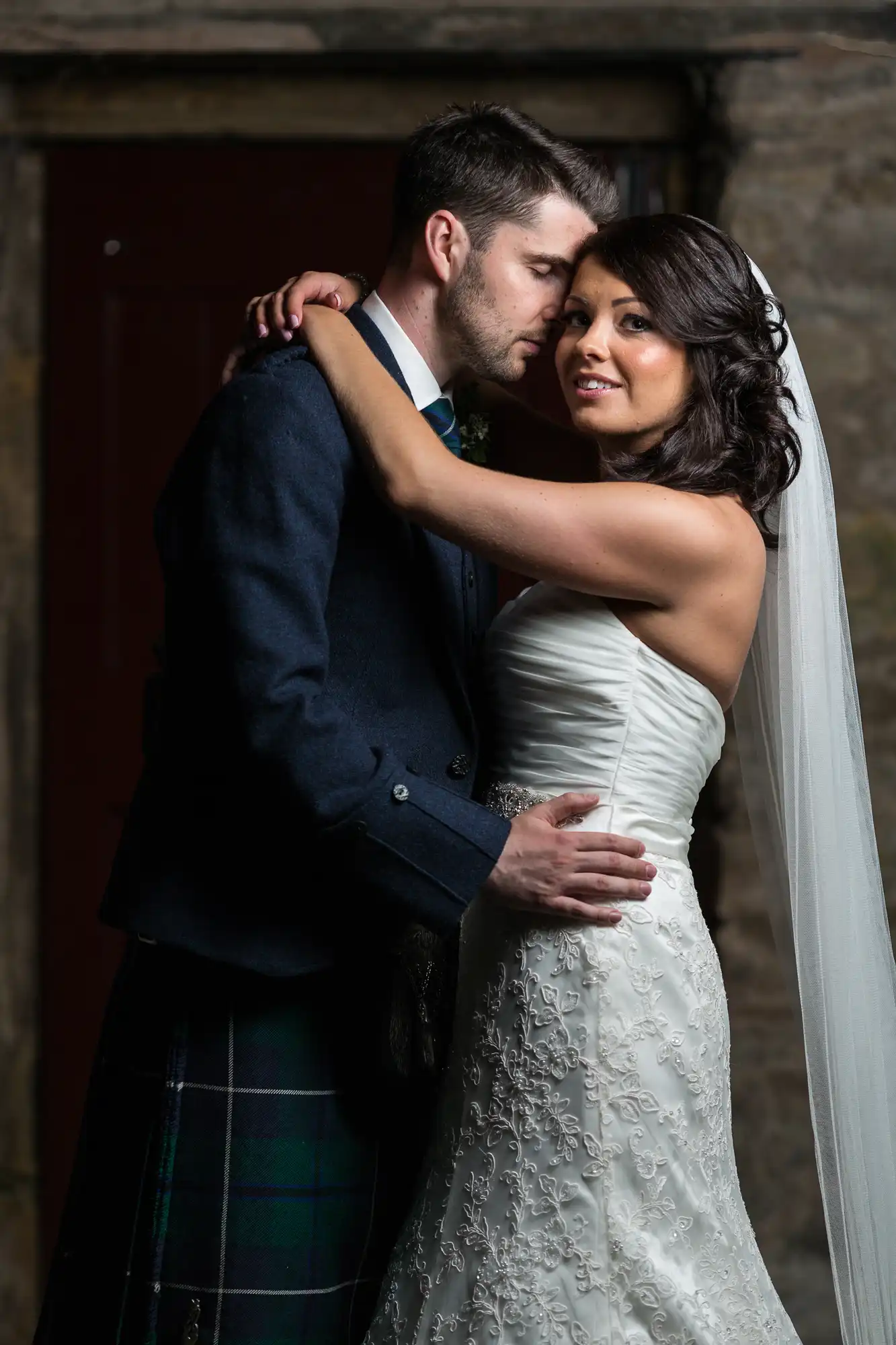 A bride and groom embrace tenderly, the groom in a tartan kilt and the bride in a strapless lace gown, with a soft focus on a rustic door background.