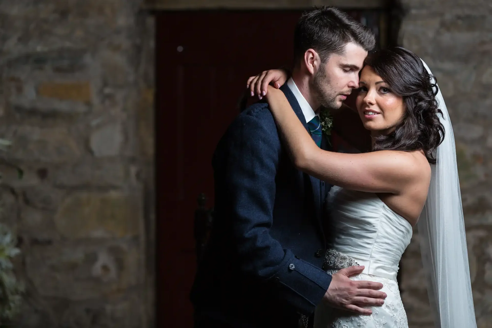 A bride and groom embrace lovingly, illuminated by soft light against a dark stone wall backdrop.