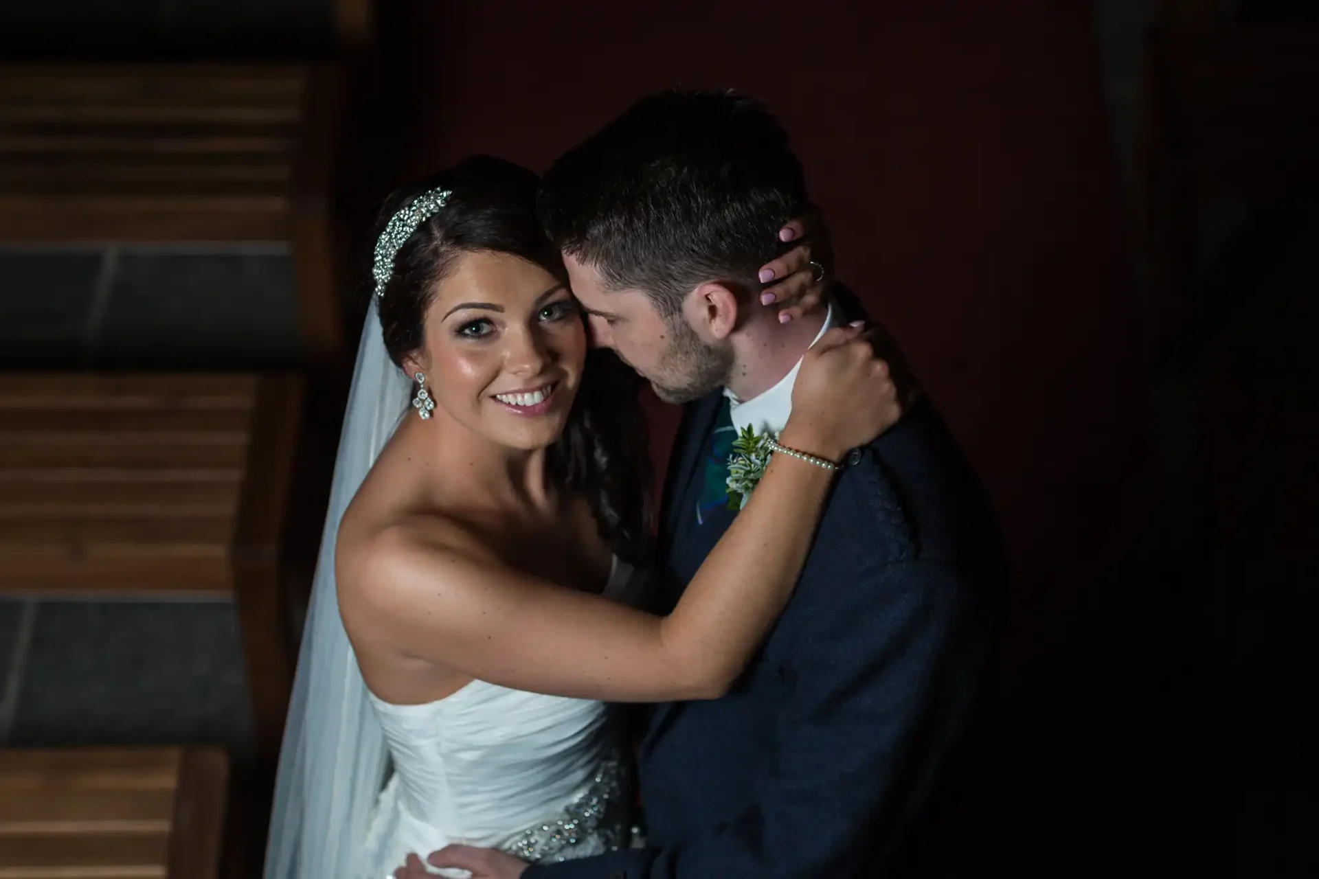 Bride in a white dress and groom in a dark suit embracing on a staircase, softly illuminated from above.