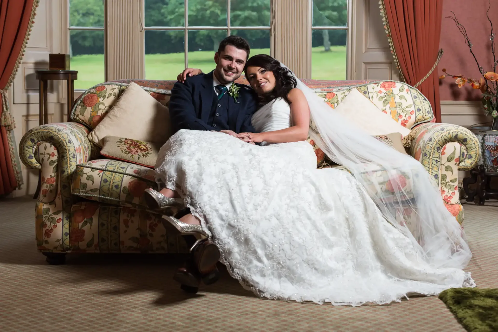 A newlywed couple smiling, seated on a floral sofa in an elegant room, the bride in a white lace gown and the groom in a dark suit with a green boutonniere.