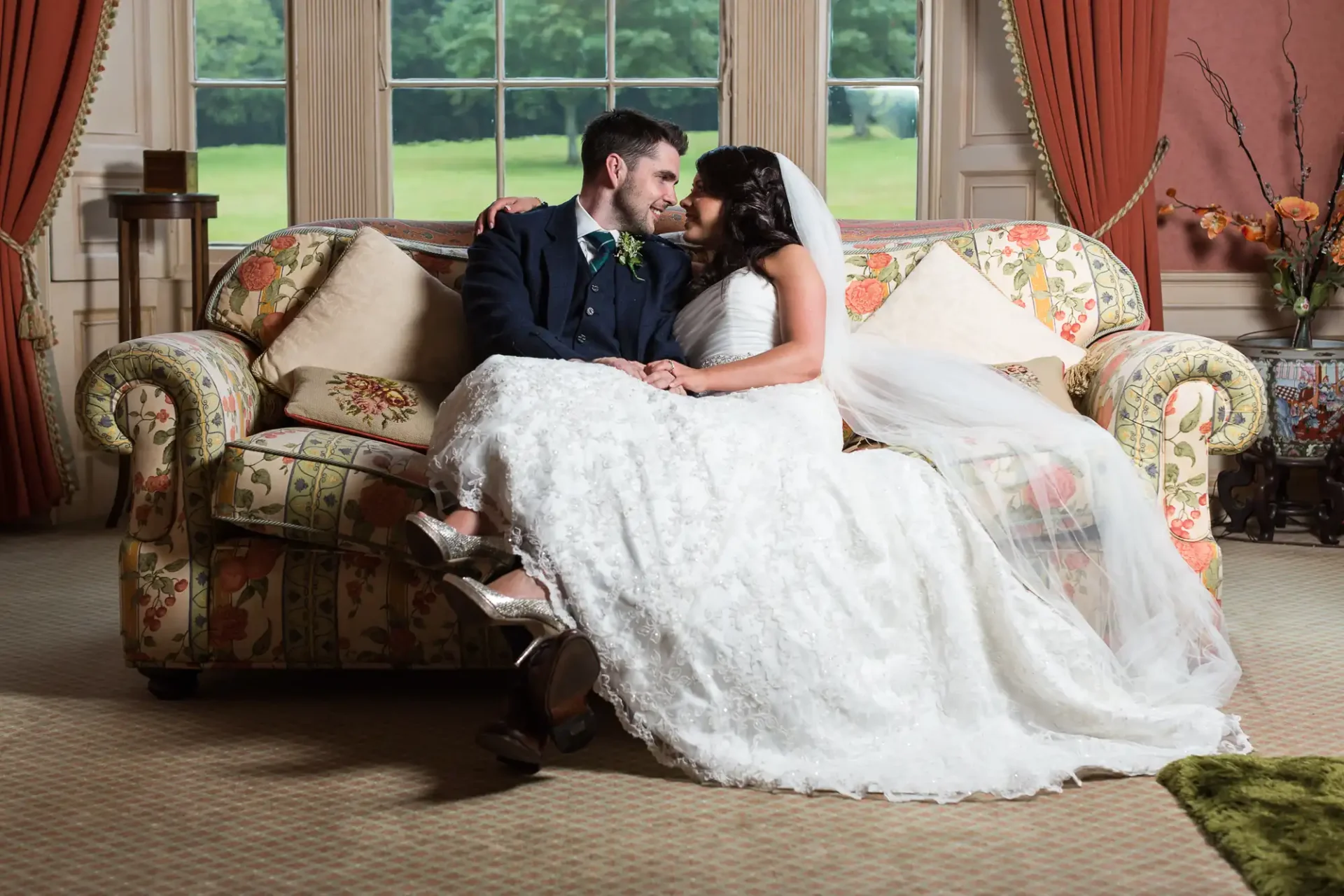 A newlywed couple sitting on a couch, smiling at each other in an elegant room, with the bride in a white dress and the groom in a navy suit.