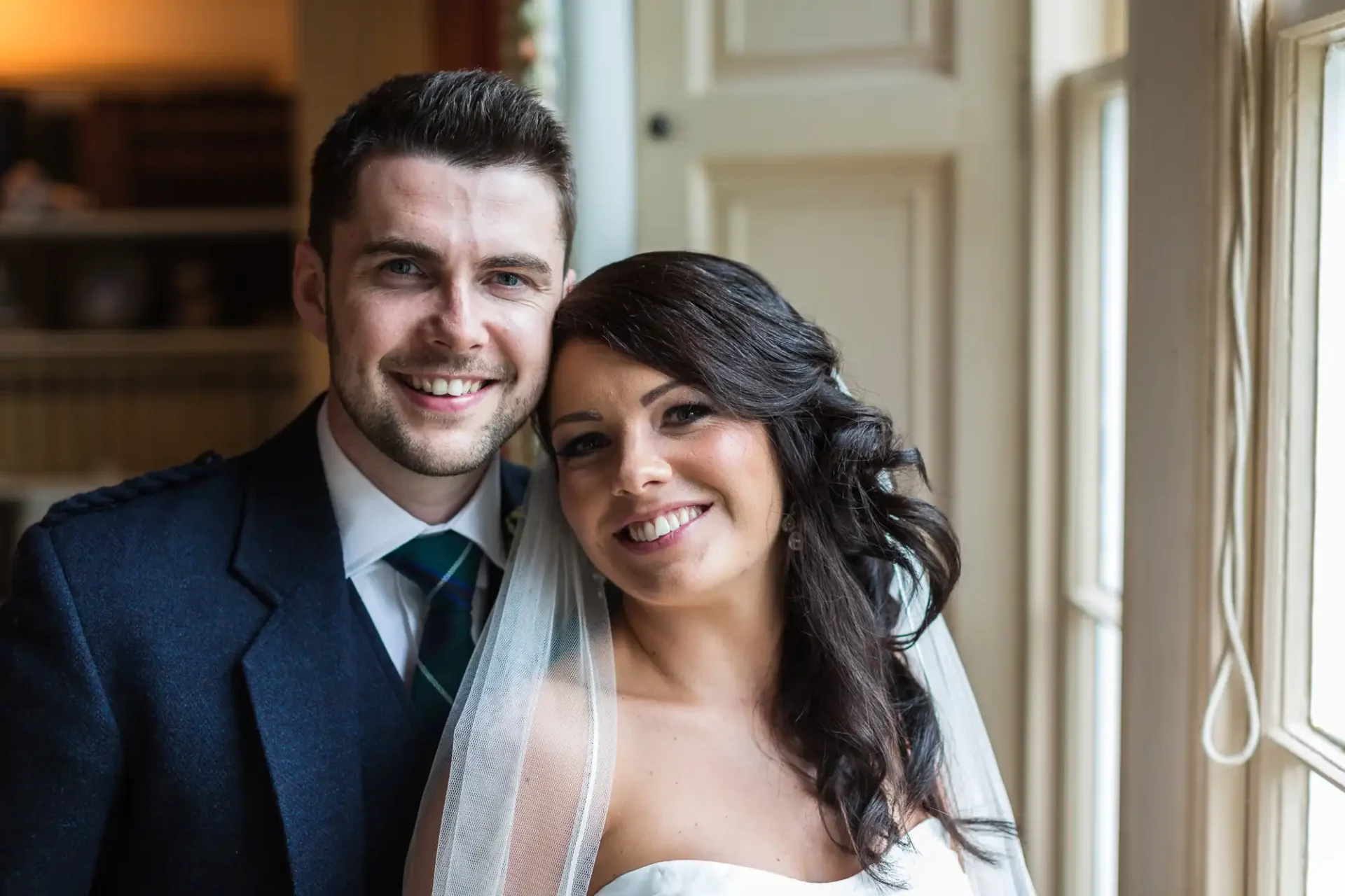 A bride and groom smiling near a window, the bride in a white dress and veil, the groom in a dark suit and tie.