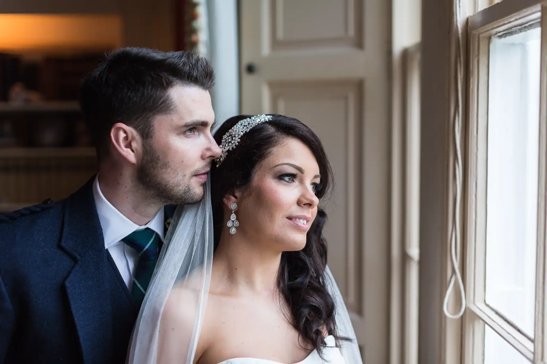 A bride and groom in formal wear gaze out a window, showing profiles with a focus on their expressions of anticipation or reflection.