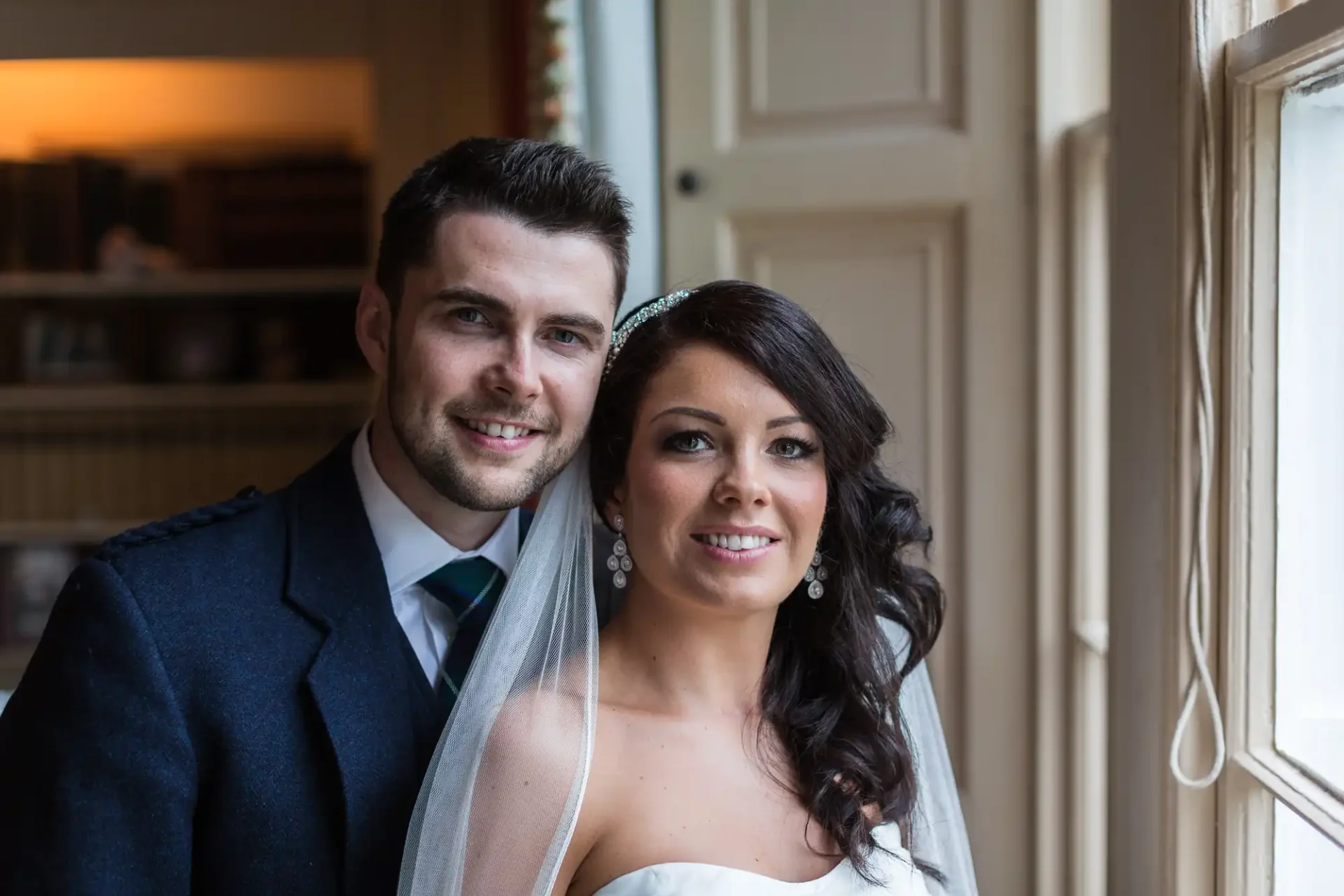A bride and groom smiling at the camera, standing by a window in a well-lit room, the bride in a white dress and veil, the groom in a dark suit.