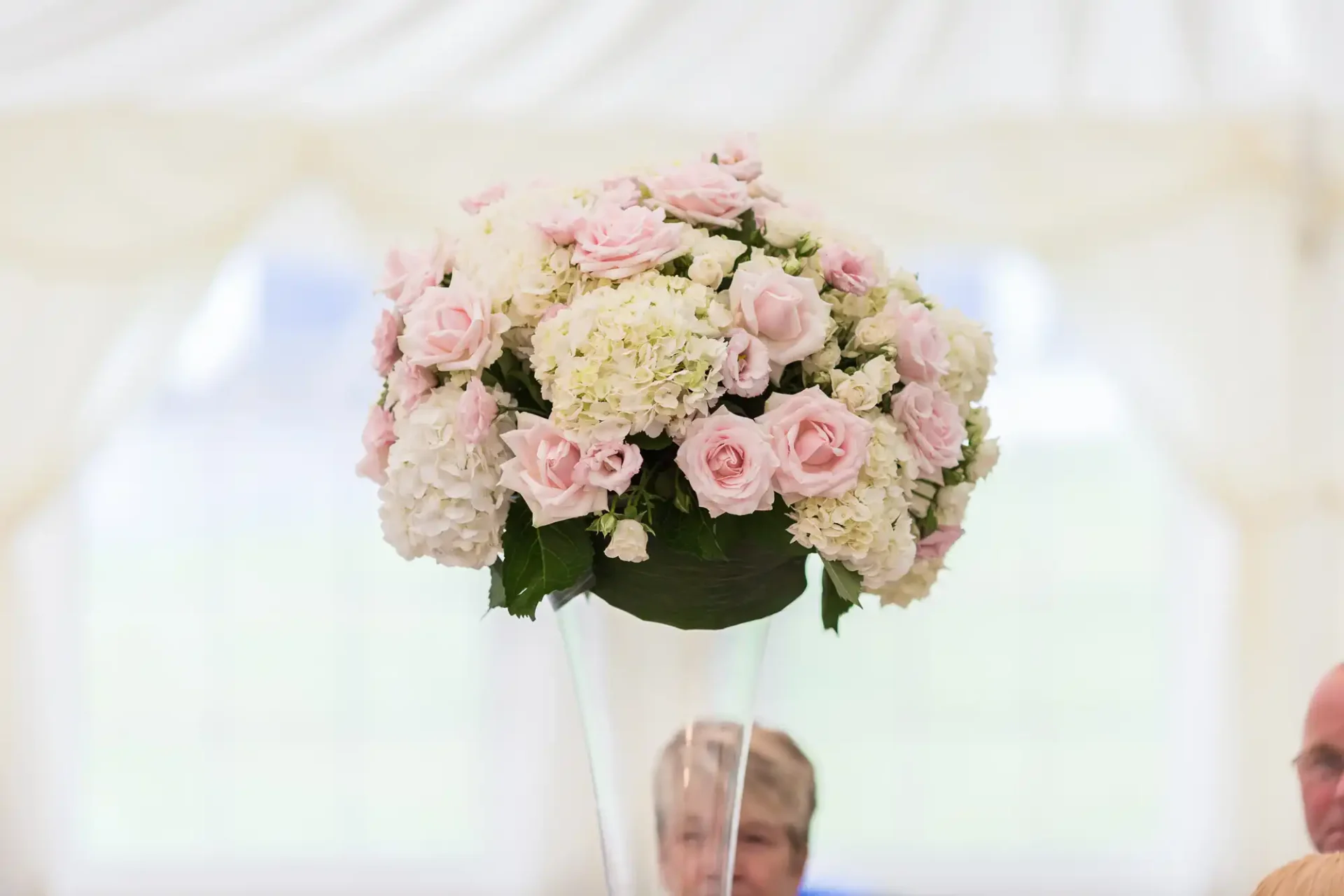 A bouquet of pink roses and white hydrangeas in a vase on a table, with people blurred in the background under a white tent.