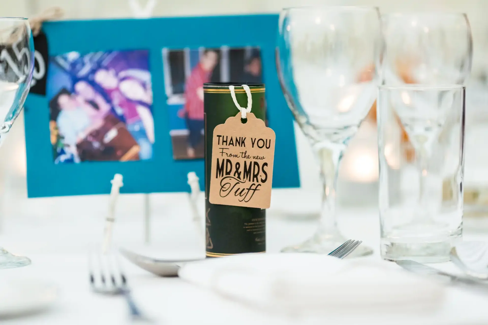 A thank you card rests on a decorated table amidst wine glasses and cutlery with a blurred photo in the background.