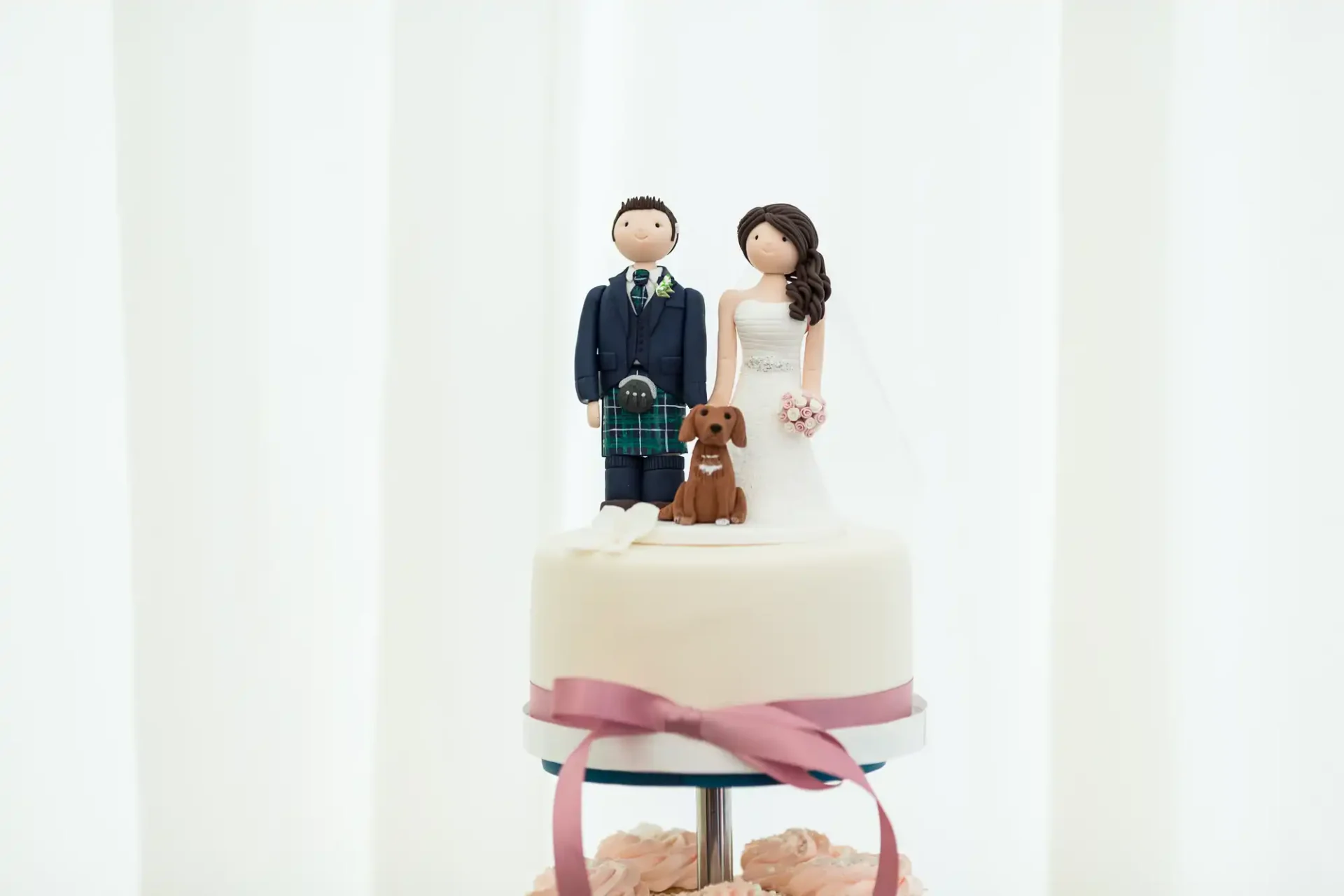 Wedding cake topper featuring figurines of a bride, groom in a kilt, and a dog, on a white cake with a pink ribbon.