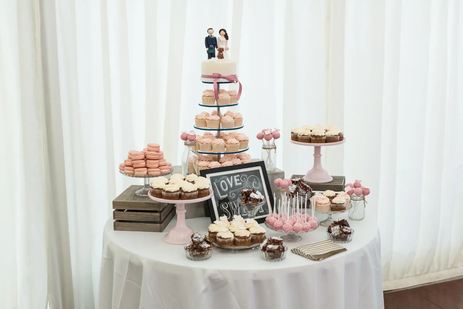 A wedding dessert table with macarons, cupcakes, and a cake topped with a figurine of a couple, set against a backdrop of white curtains.