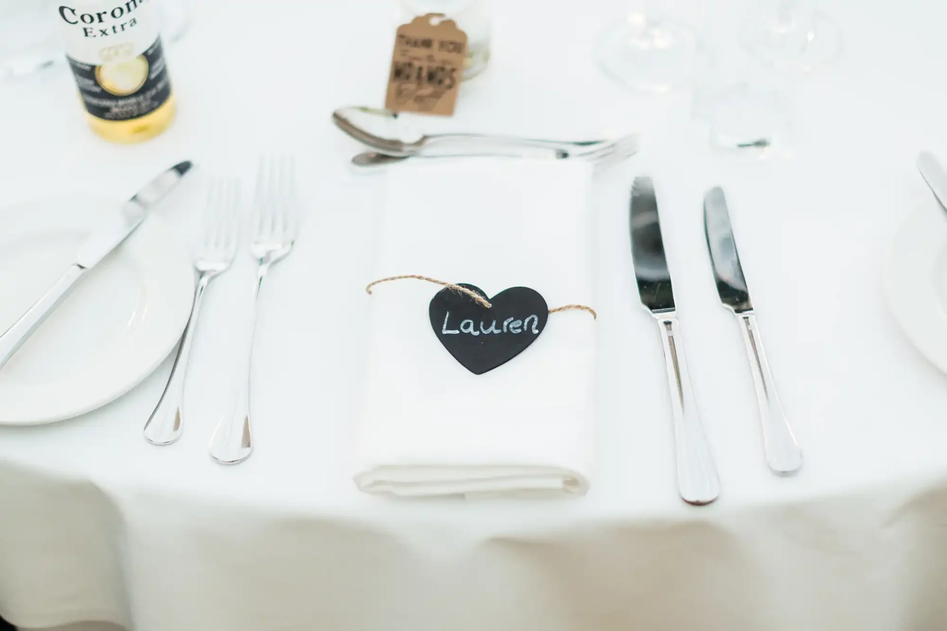 Elegant dining table setting with a white napkin, silver cutlery, and a name tag shaped like a heart reading "lauren" on a white tablecloth.